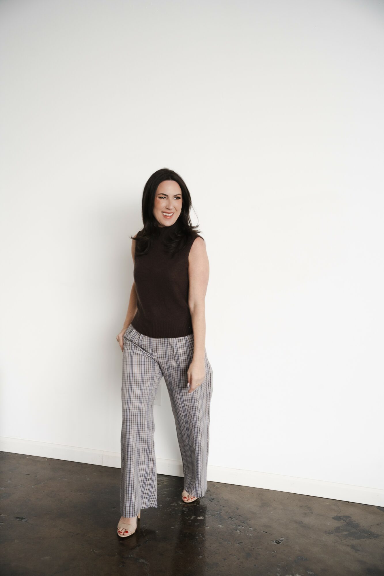 Woman standing against a plain white wall, smiling, wearing a sleeveless dark top and striped pants.