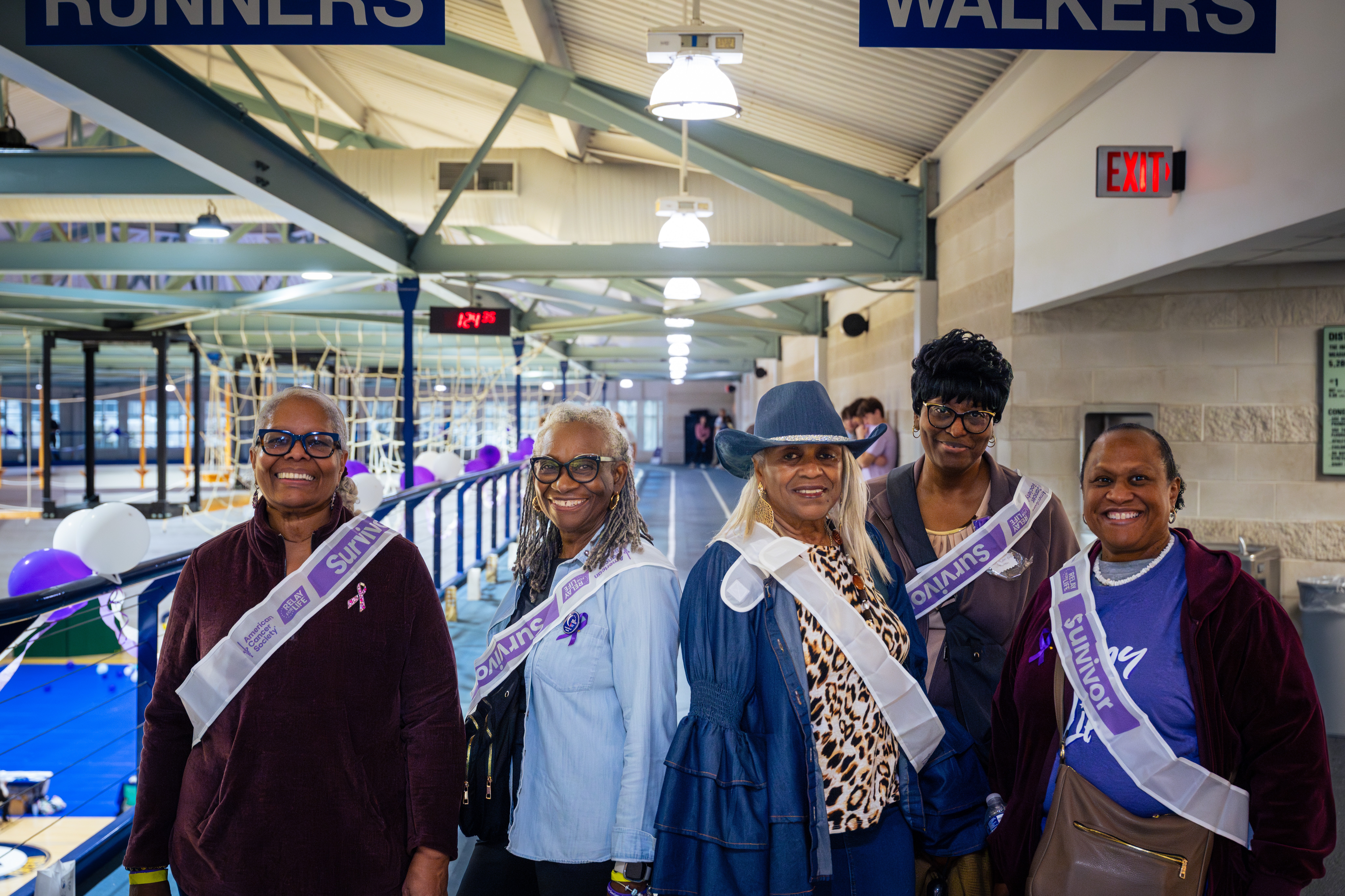 Five women standing together indoors, wearing sashes and smiling, with a roller coaster in the background.