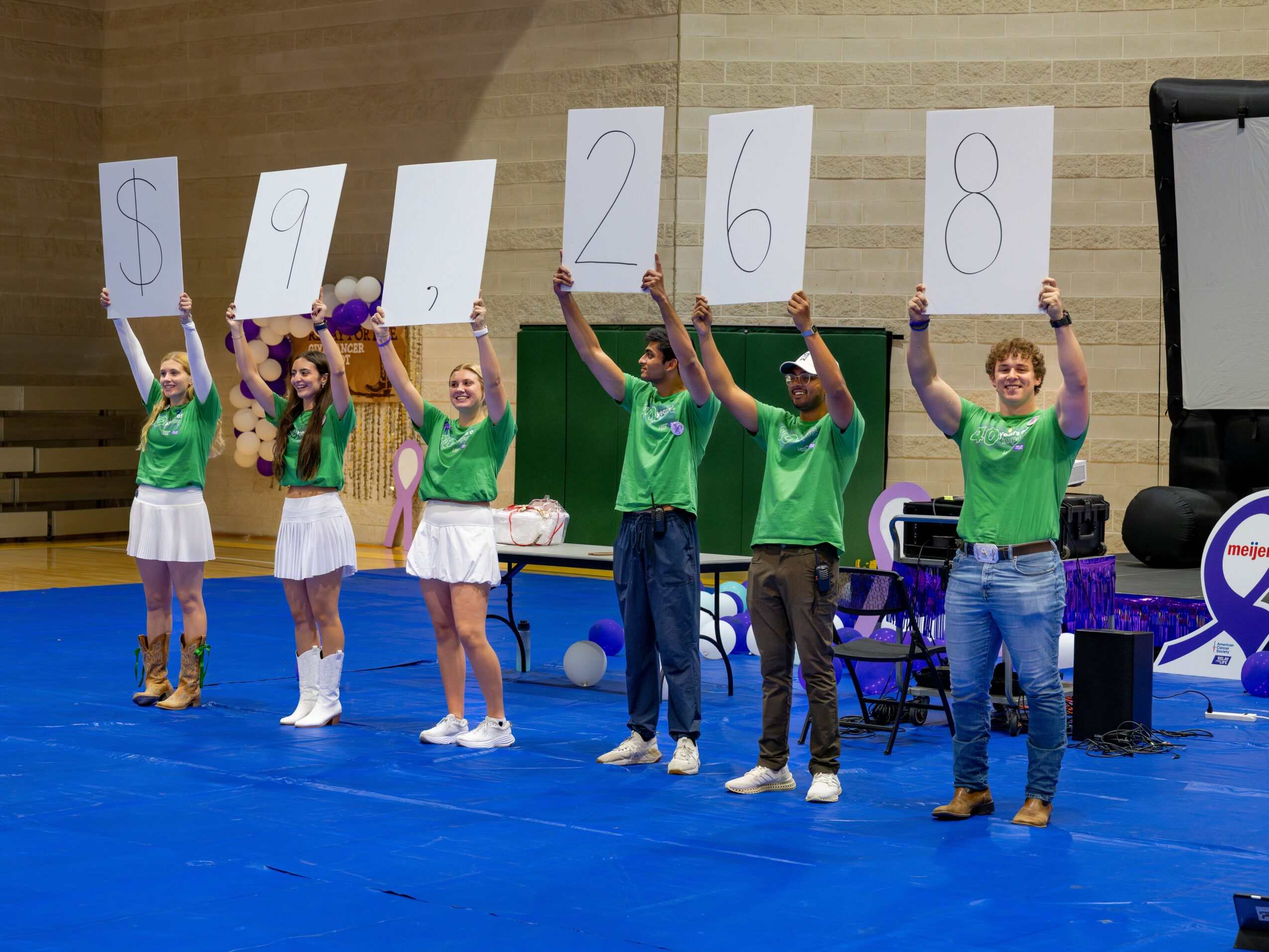 Six people on stage holding large signs with numbers and a dollar sign, wearing green shirts and white skirts or pants, in a gymnasium.