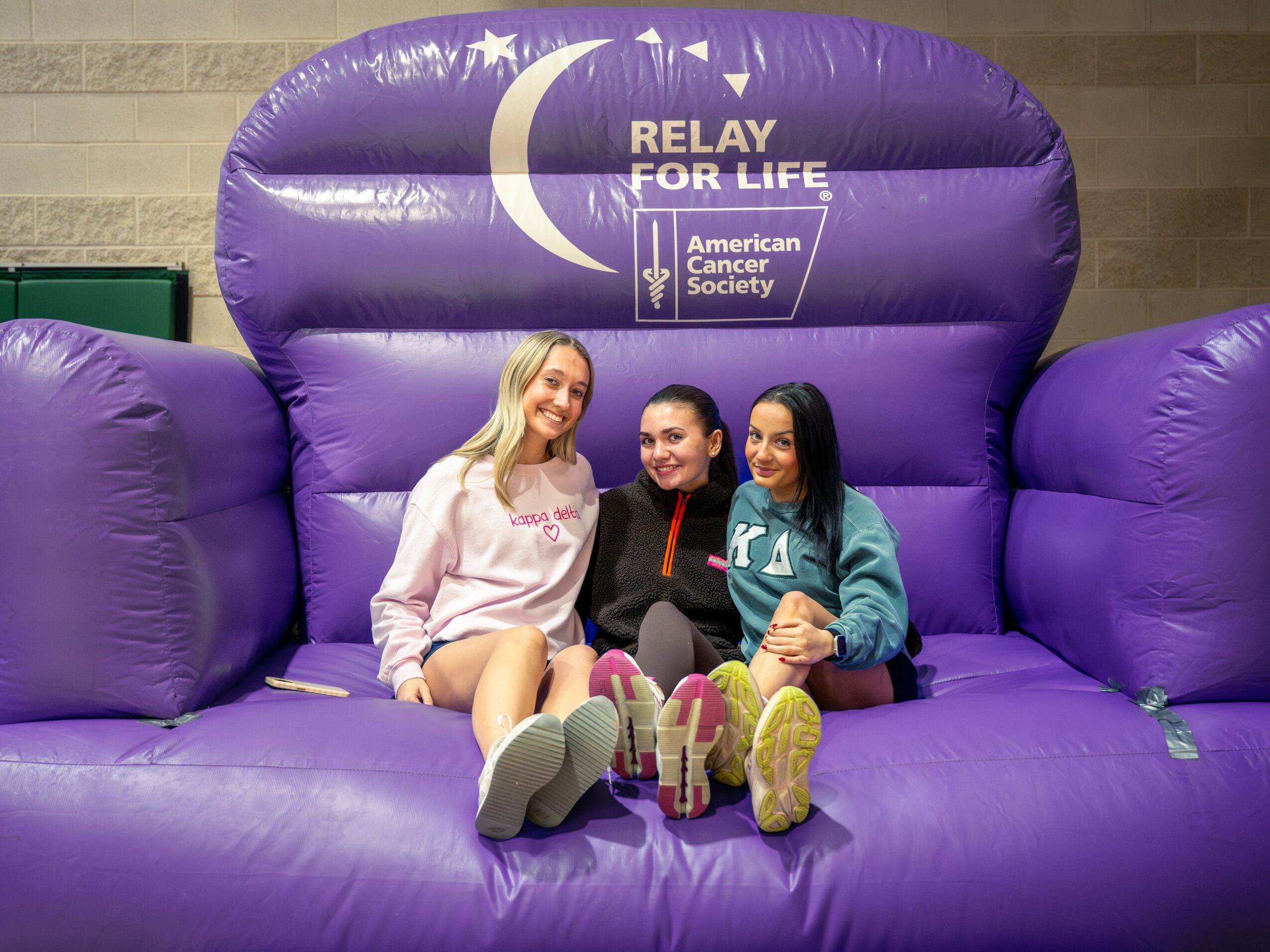Three women sitting on a large purple inflatable with a crescent moon and text, smiling at the camera.