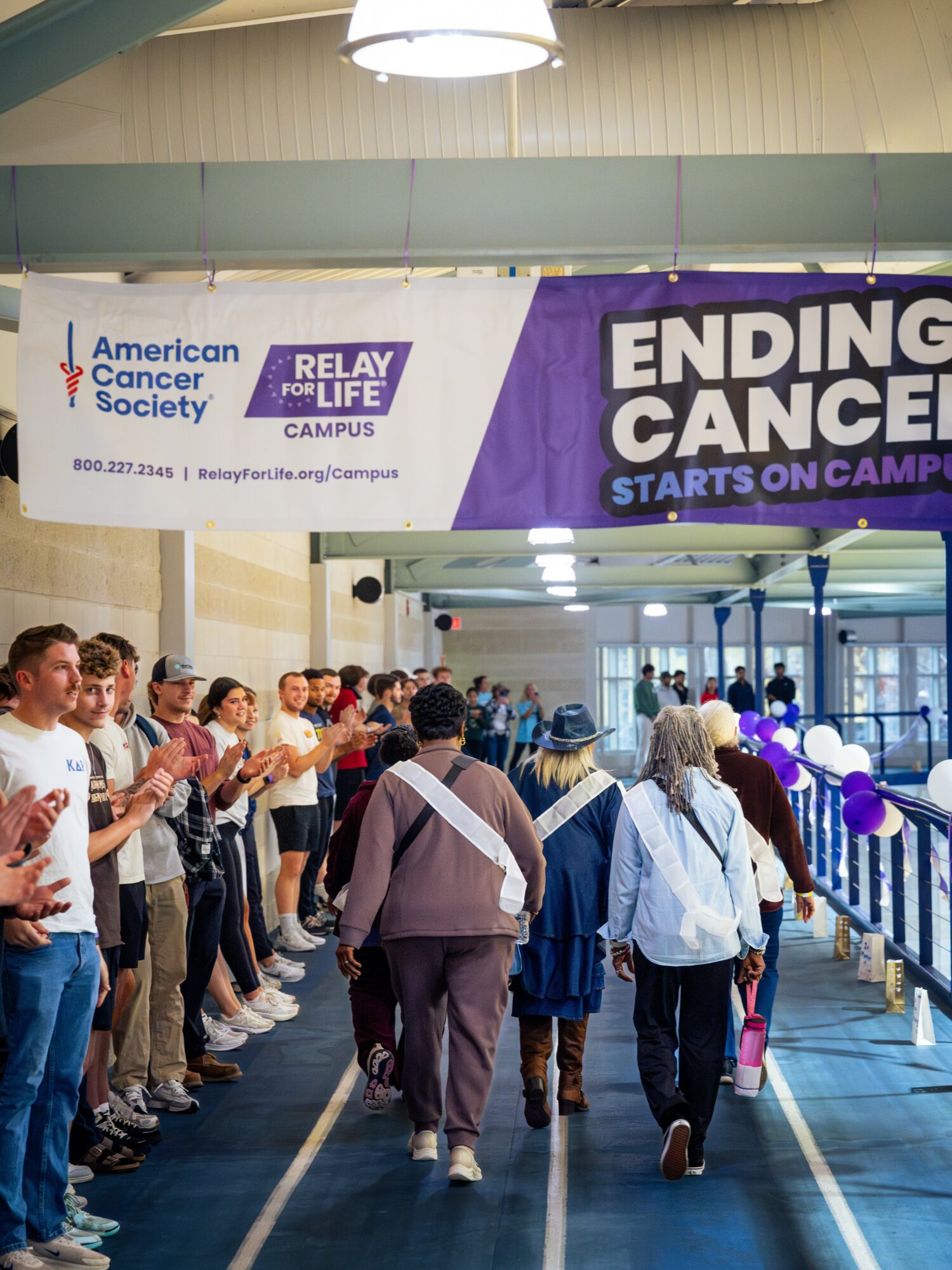 People walking through a cancer awareness event with a banner reading 'Ending Cancer Starts on Campus' and others clapping nearby.