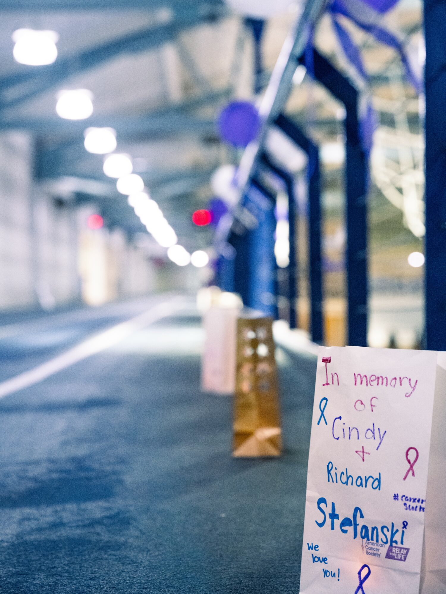 Memorial on sidewalk with bags, pink ribbon, and handwritten note in memory of Cindy Richard Stefanski, with blue and pink text.