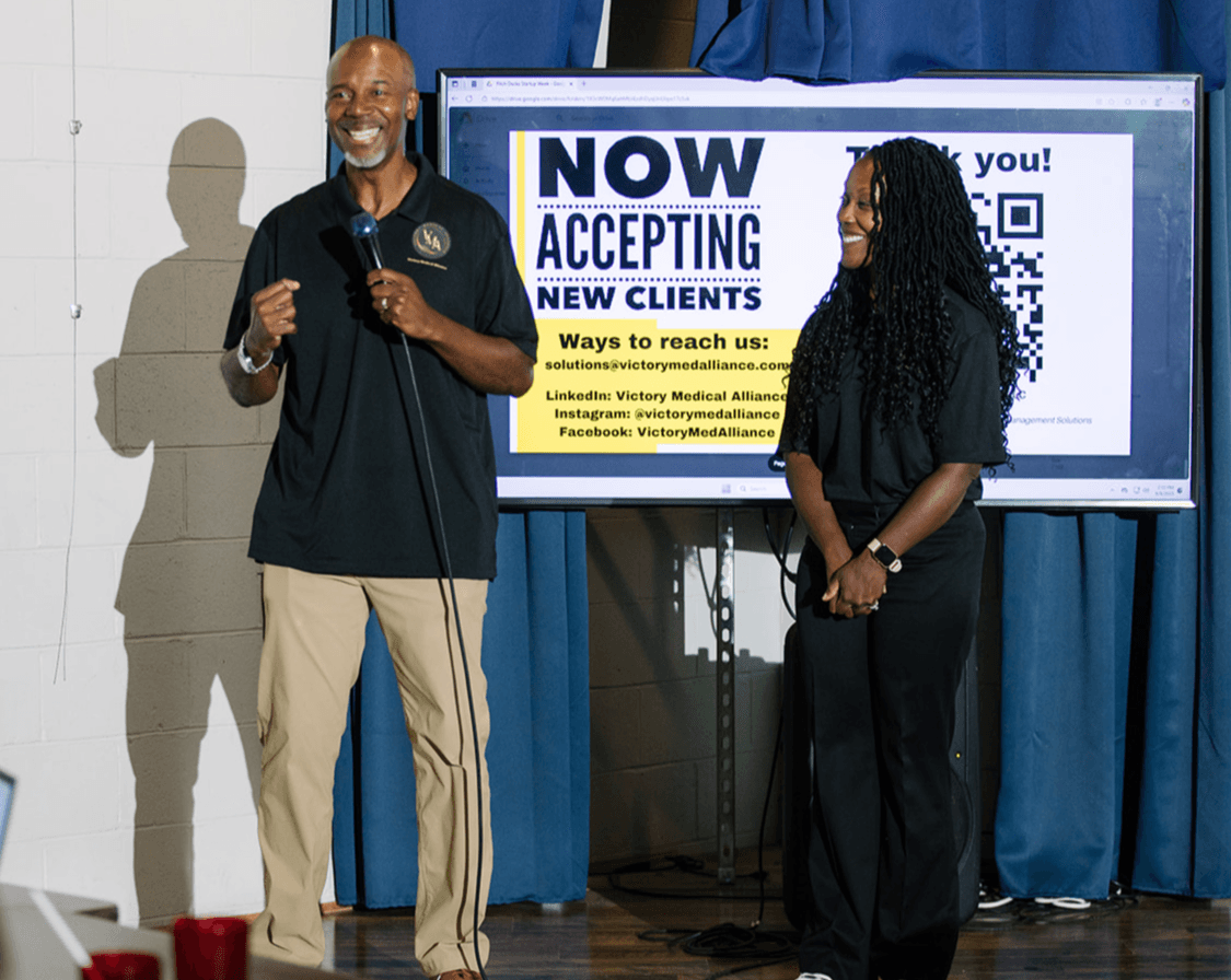 Two people standing on stage, one speaking into a microphone, the other smiling, with a large sign behind them.