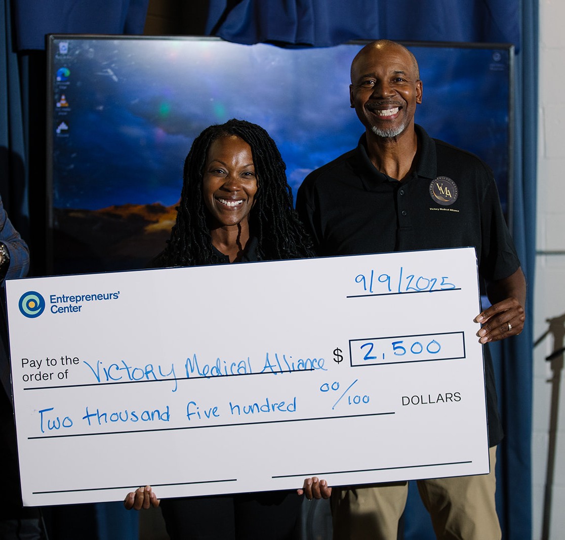 Two people holding a large check for $2,500 made out to Victory Medical Alliance, date 9/9/2022, in front of a screen.