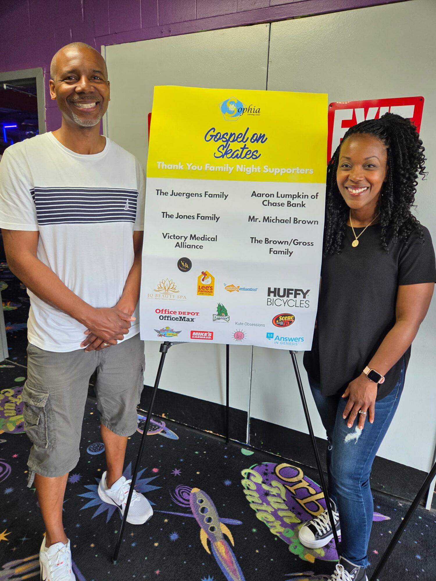 Two smiling people standing next to a colorful poster on an easel, in an indoor setting with a dark carpeted floor.