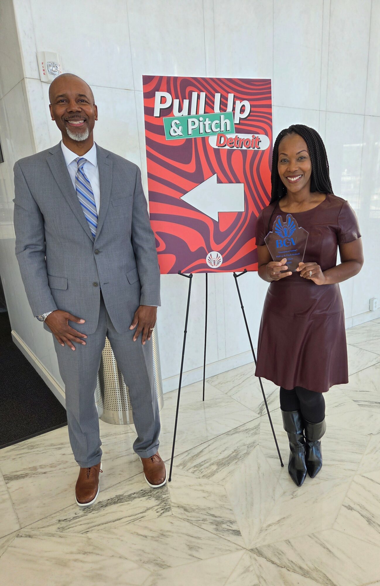 A man in a gray suit and a woman in a maroon dress stand next to a sign with an arrow pointing left, smiling.