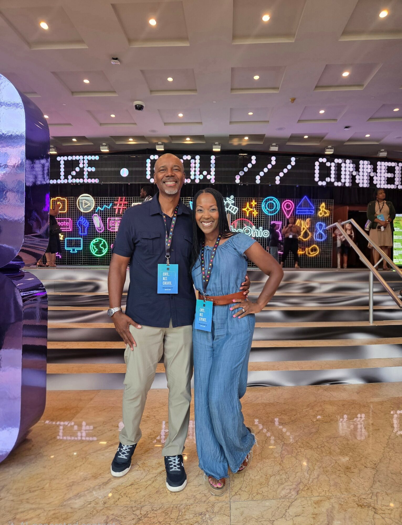 Two people smiling and standing together in front of a digital display at an event, with stairs behind them.