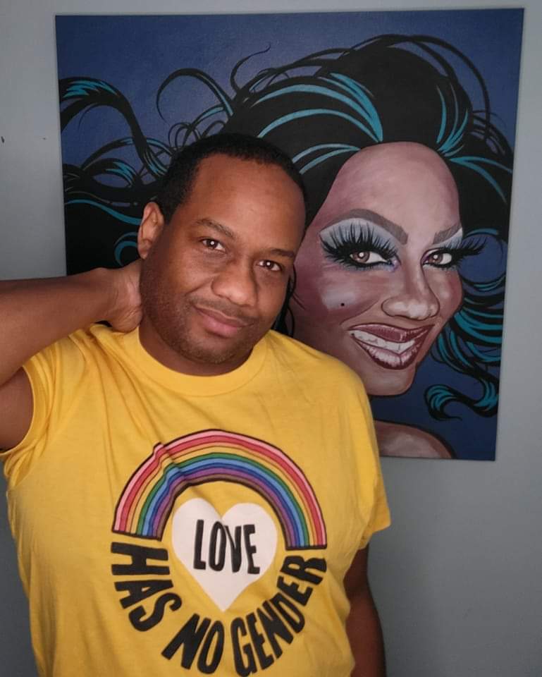 Man in yellow shirt standing in front of colorful portrait painting, smiling, with hand on neck.