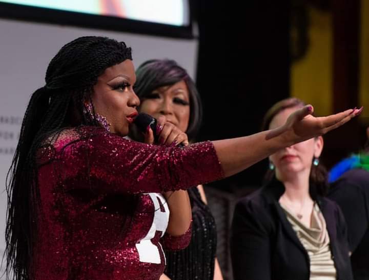 Woman speaking into microphone, gesturing with her hand, others listening on stage, dark background, formal event, three women visible.