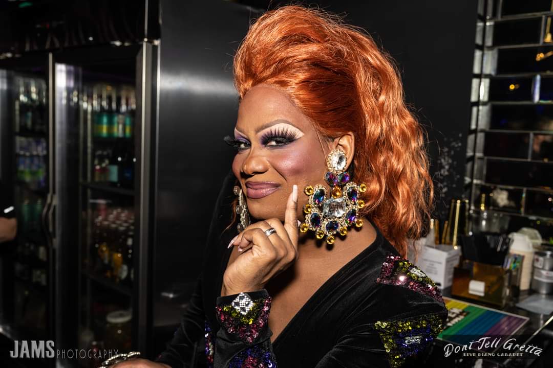 Person with orange hair, makeup, and large earrings, smiling and touching chin, in a dark room with shelves and bottles