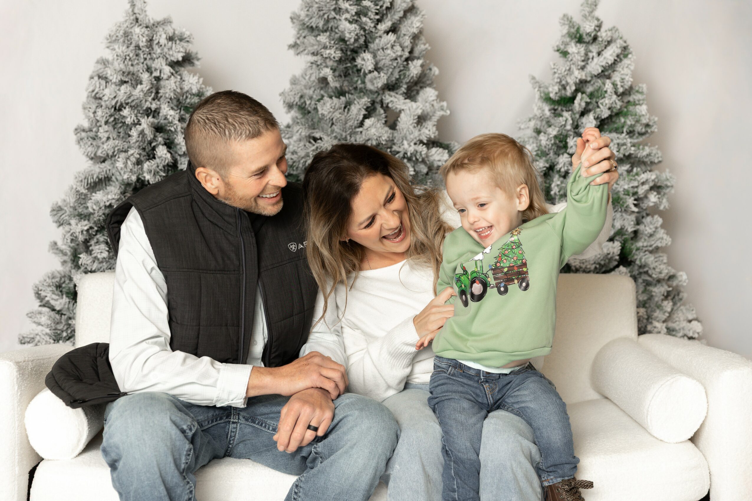 Family of three sitting on a white couch with snow-covered trees in the background, smiling and playing together.