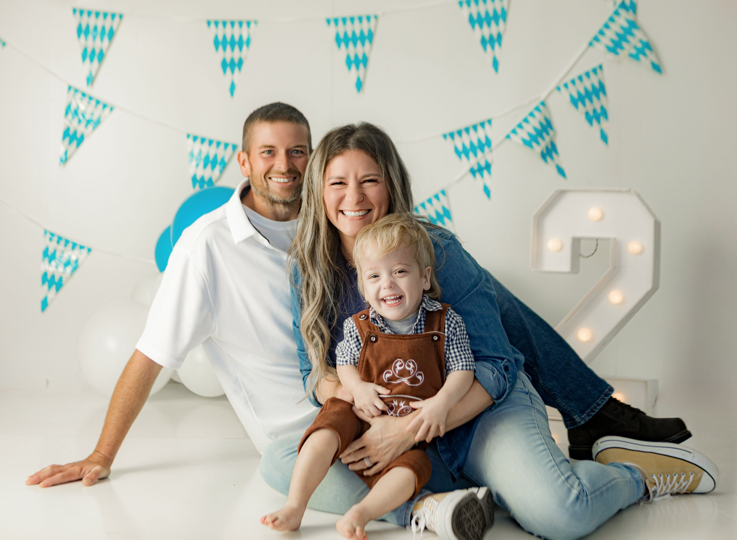 Family of three sitting together, smiling, with blue and white bunting and a large number two in the background.