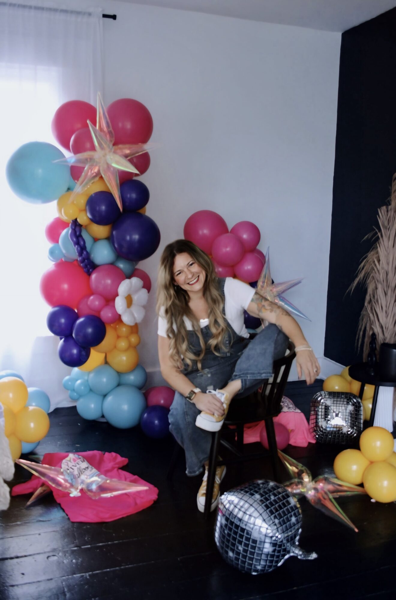 Woman sitting on a chair with colorful balloons and decorations around her, celebrating a party or event.