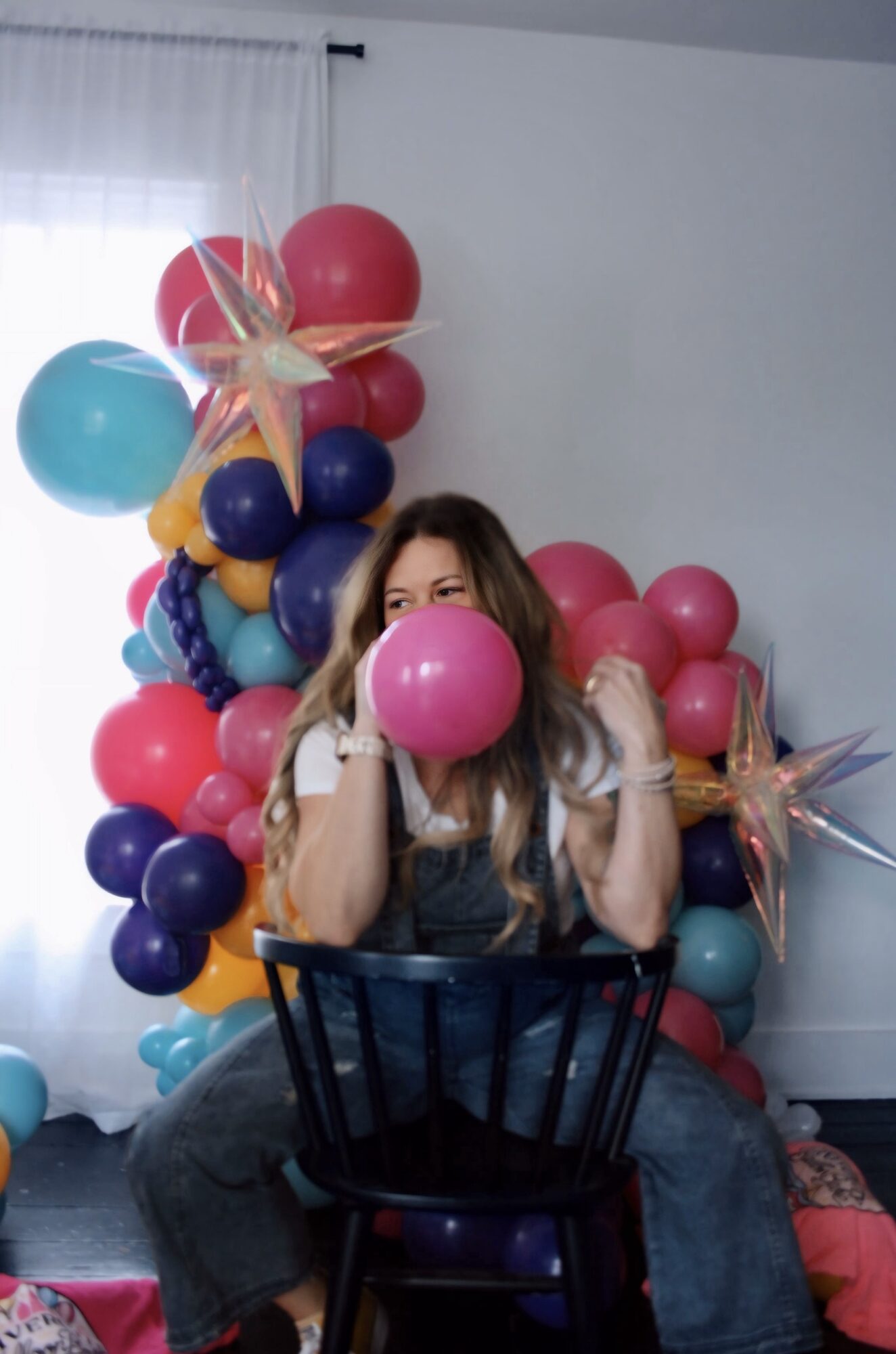 Person sitting on a chair holding a pink balloon, surrounded by colorful balloons and decorations, in a room with white walls and curtains.