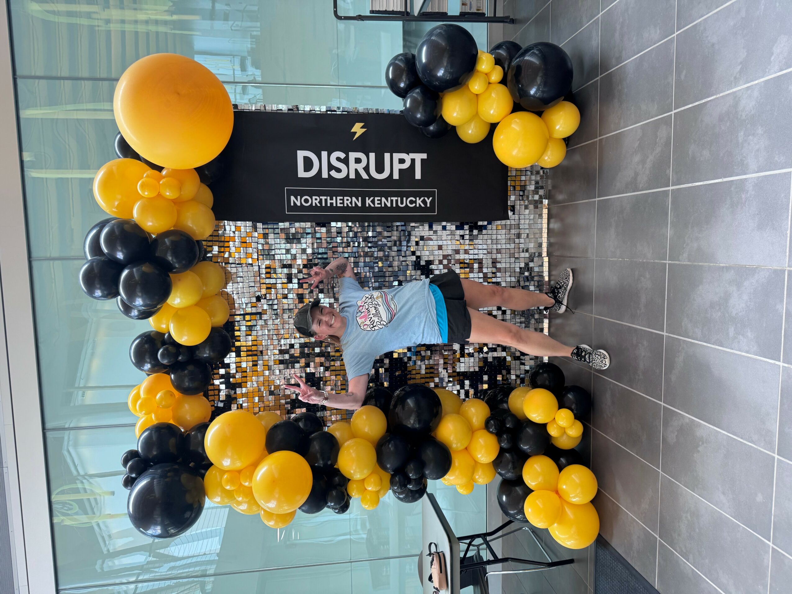 Person standing in front of a balloon arch with black and yellow balloons at an event, with a sign reading 'DISRUPT NORTHERN KENTUCKY'.