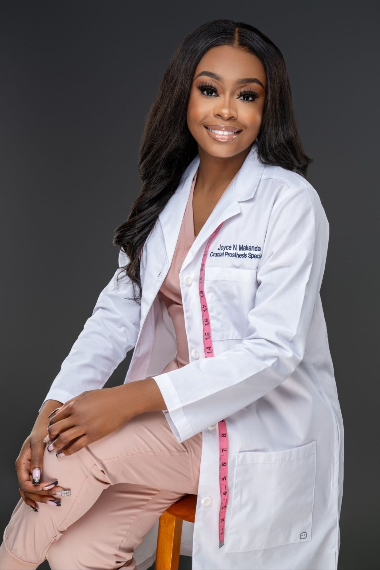 Young woman in white medical coat sitting on a stool, smiling, with a dark background.