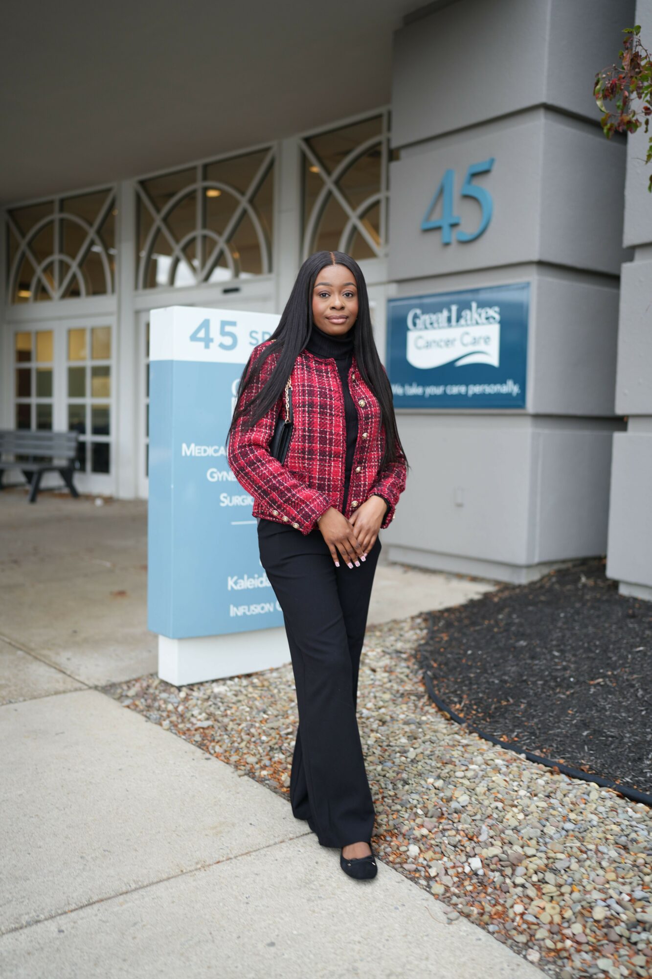 Woman standing outdoors in front of a building, wearing a red plaid shirt and black pants, smiling.