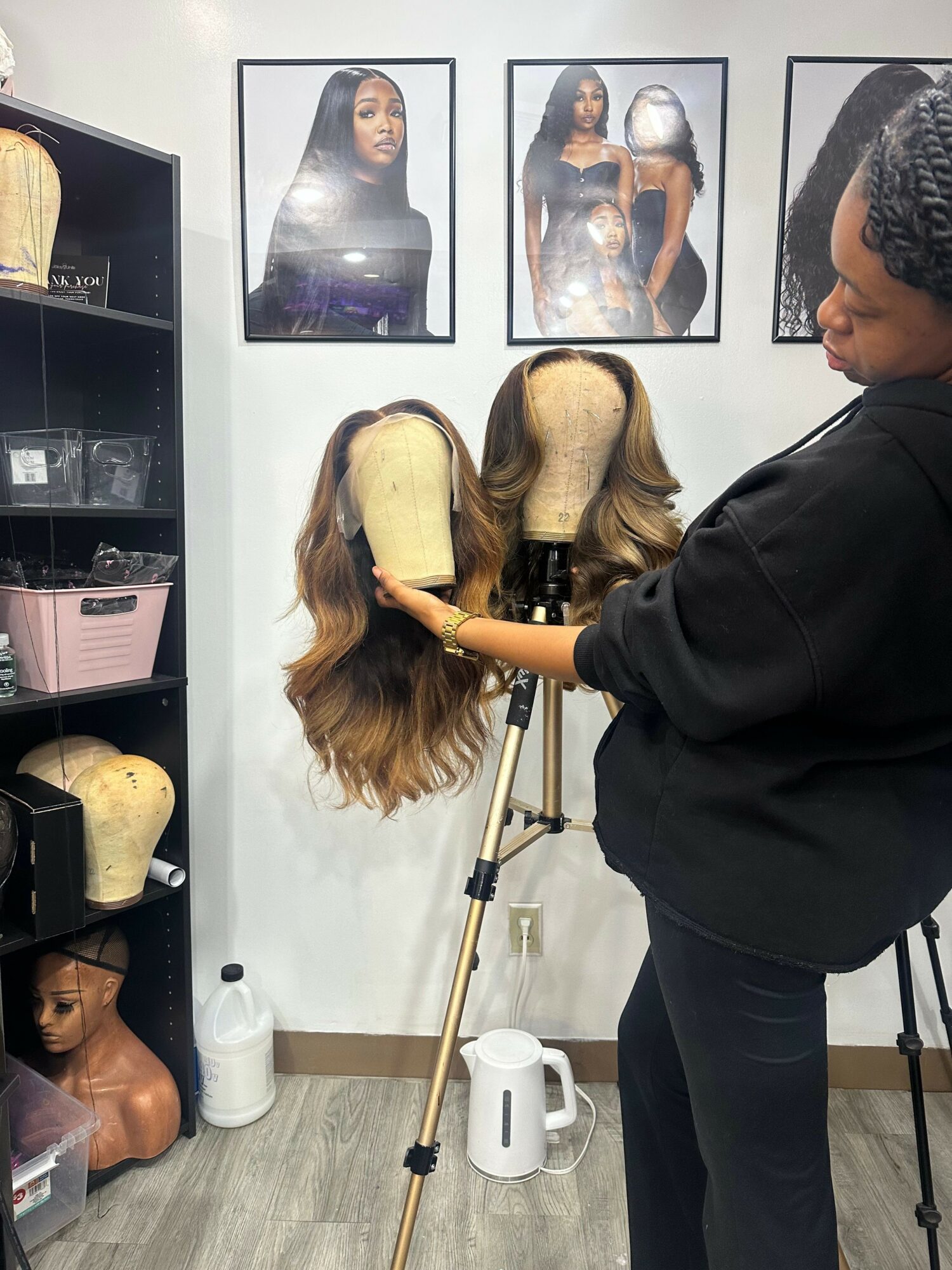 Person styling a woman's hair in a salon, with framed photos on the wall and a shelf with hair products.