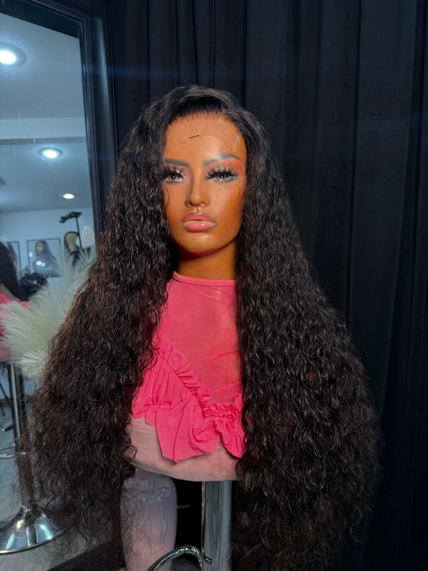 Woman with long curly hair wearing a pink top, standing indoors with dark curtains and a mirror in the background.