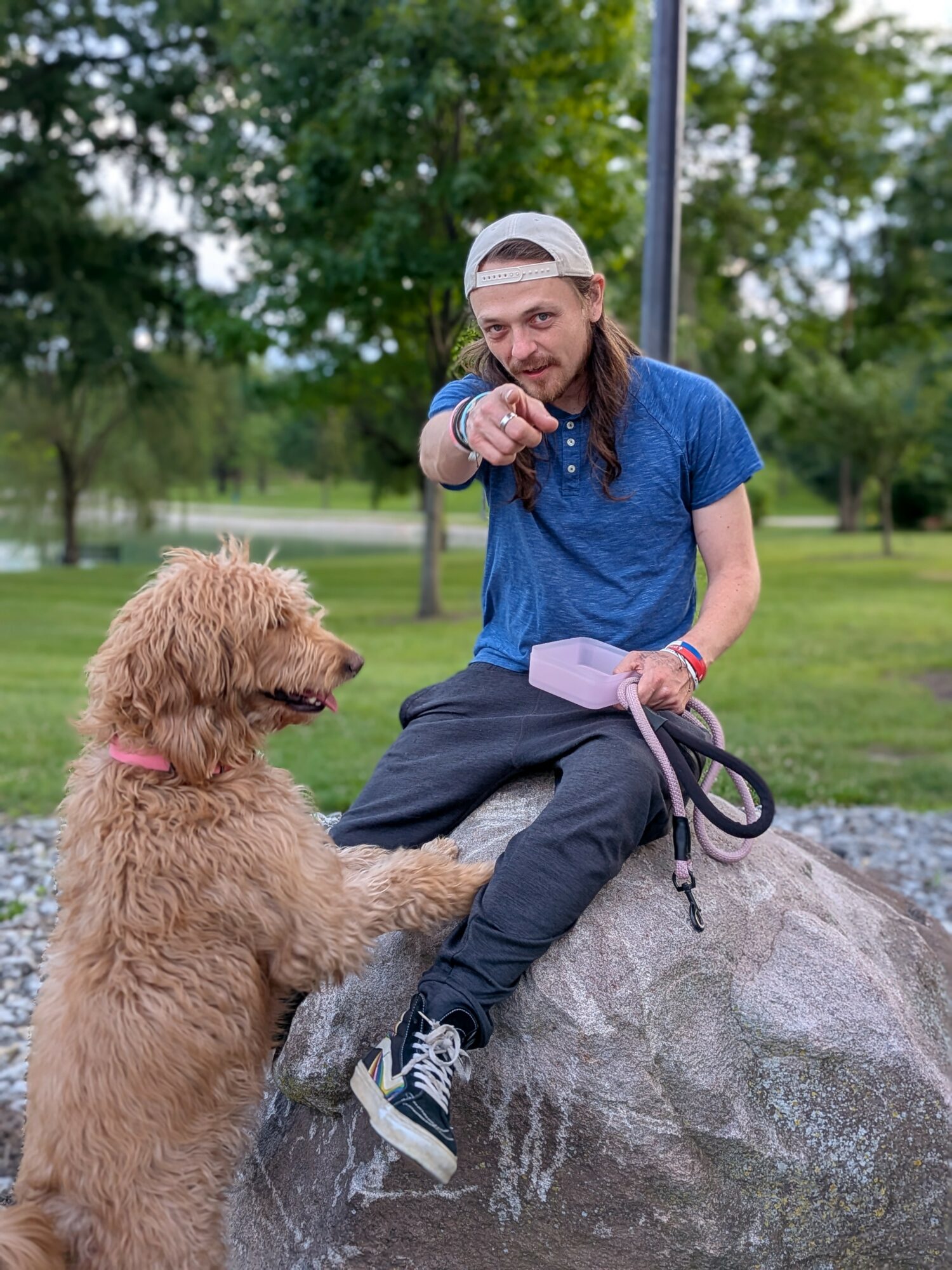 Person with long hair, wearing a blue shirt and white cap, sitting on a rock, pointing at a dog in a park.