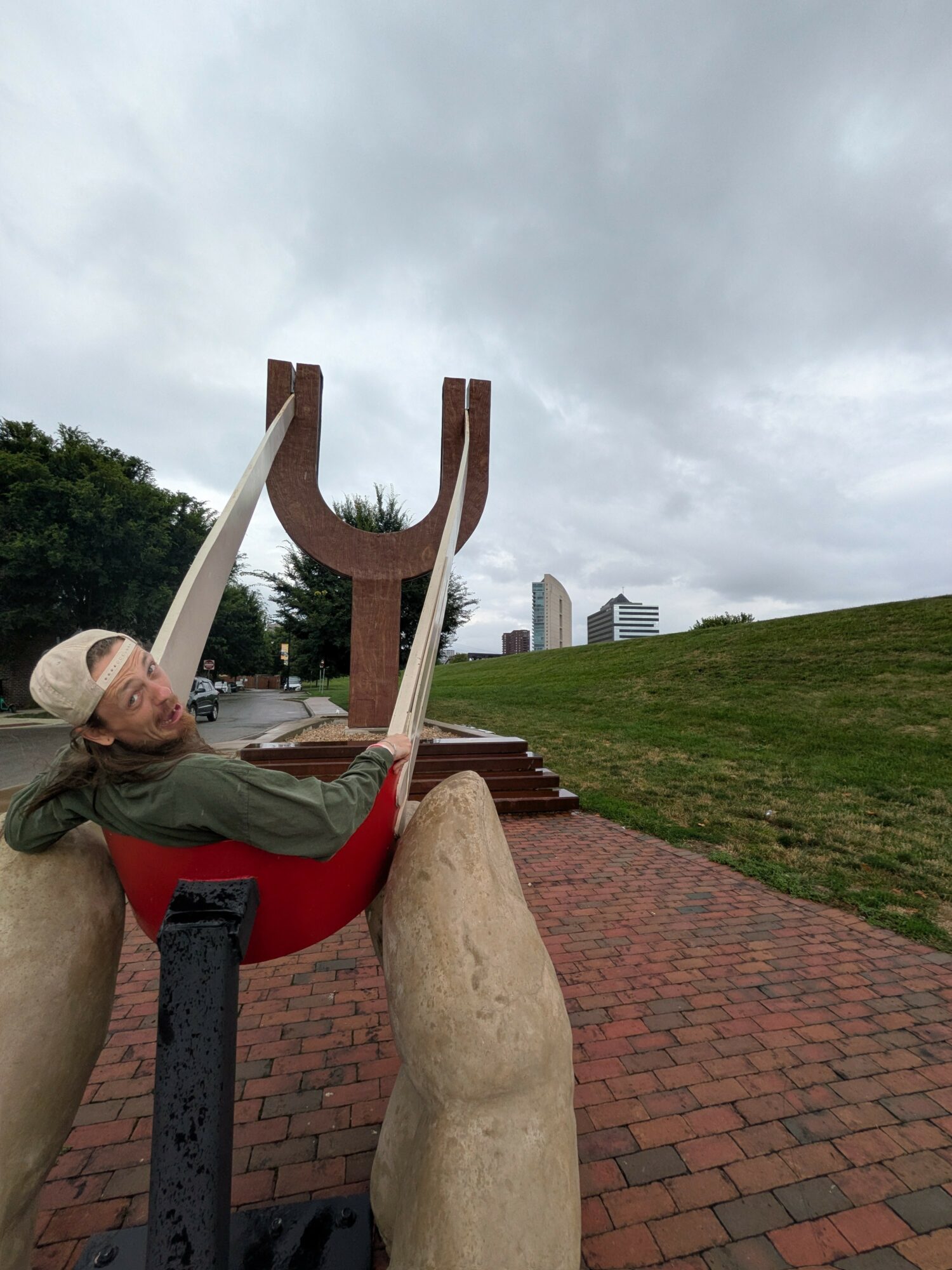Person lying on a red and black chair outdoors, with a large sculpture and buildings in the background under cloudy sky.