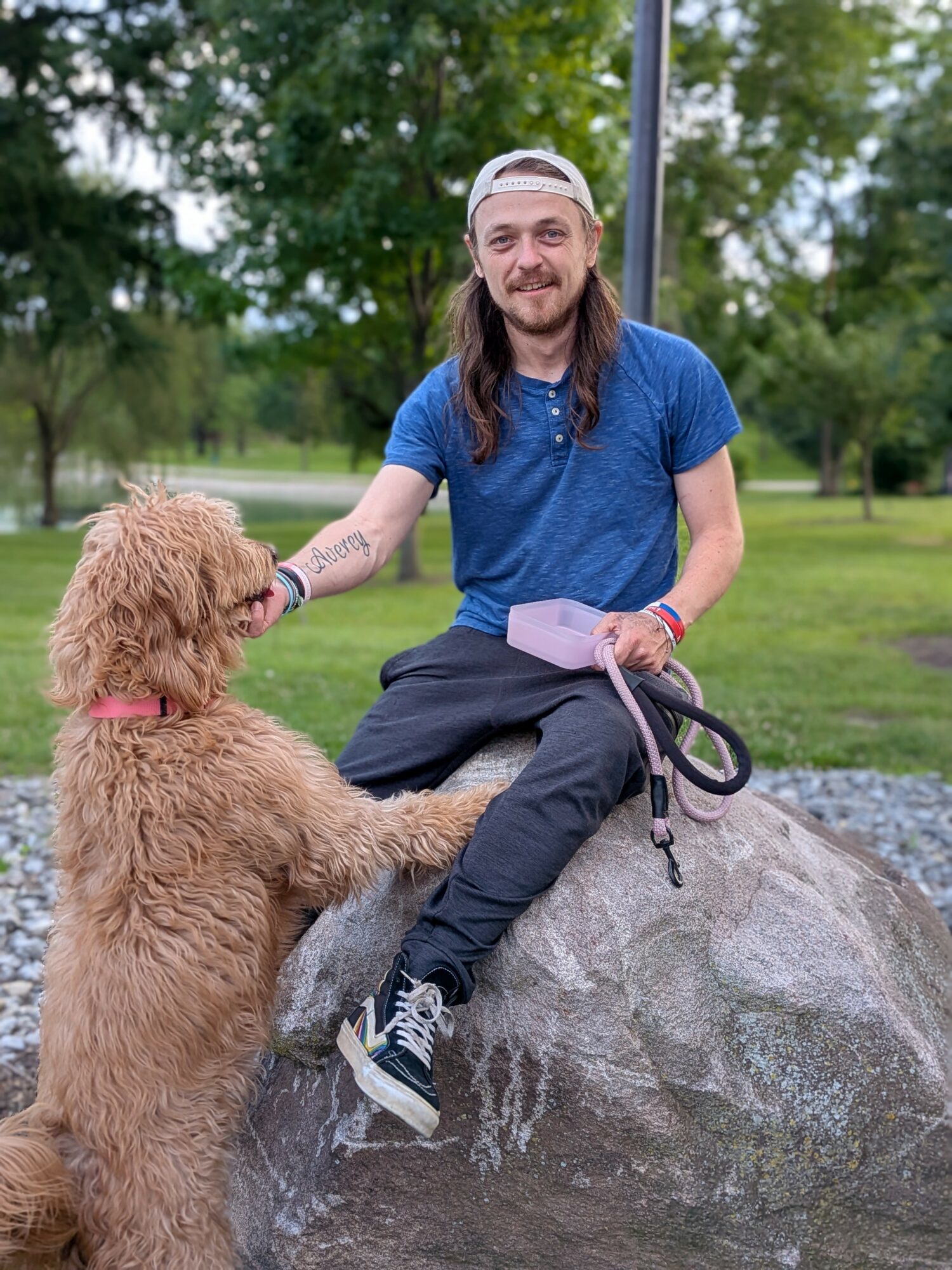 Person with long hair, wearing a blue shirt and cap, sitting on a large rock, petting a dog in a park.