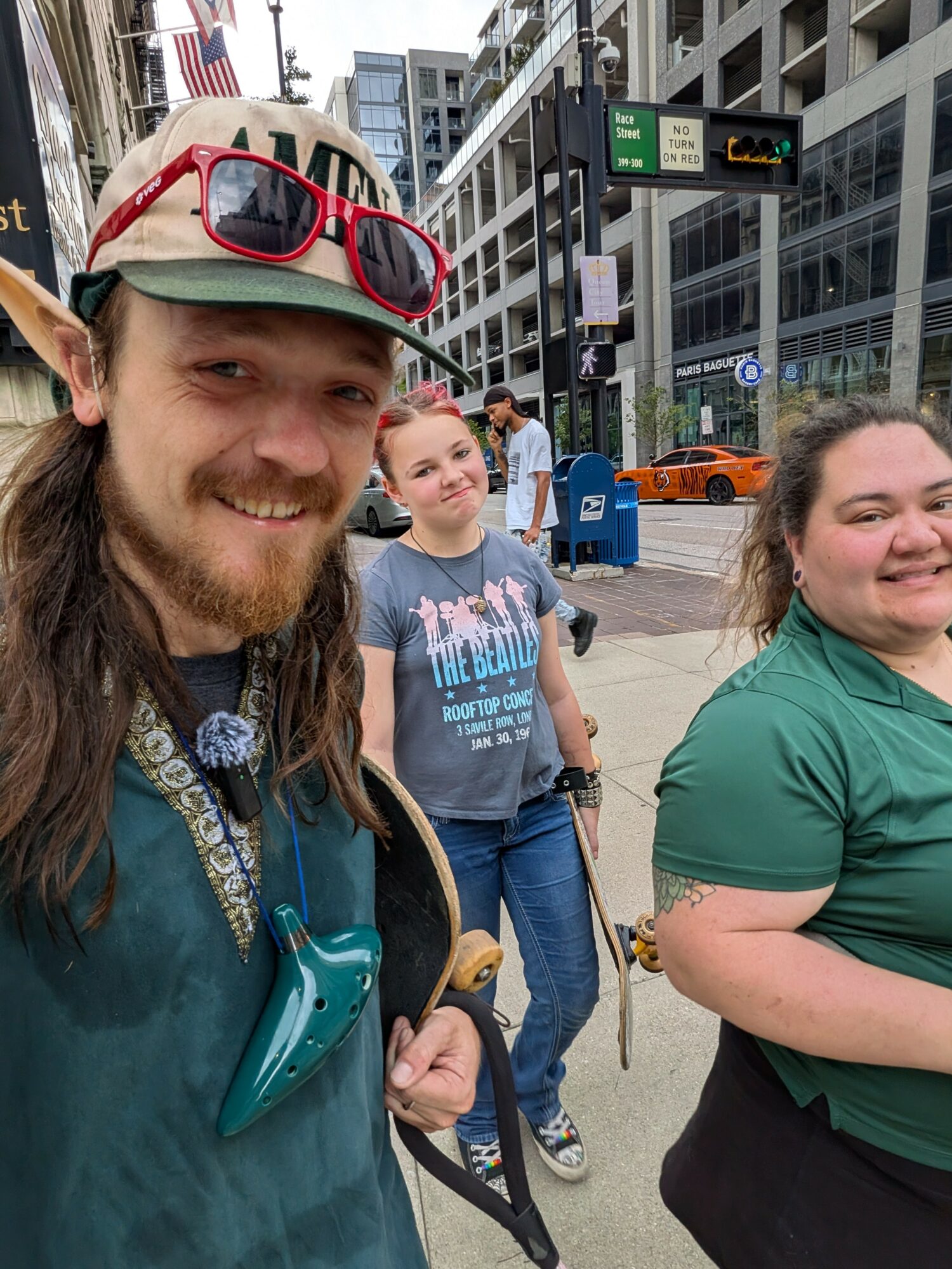 Three people walking on a city sidewalk with tall buildings, traffic lights, and a taxi in the background.