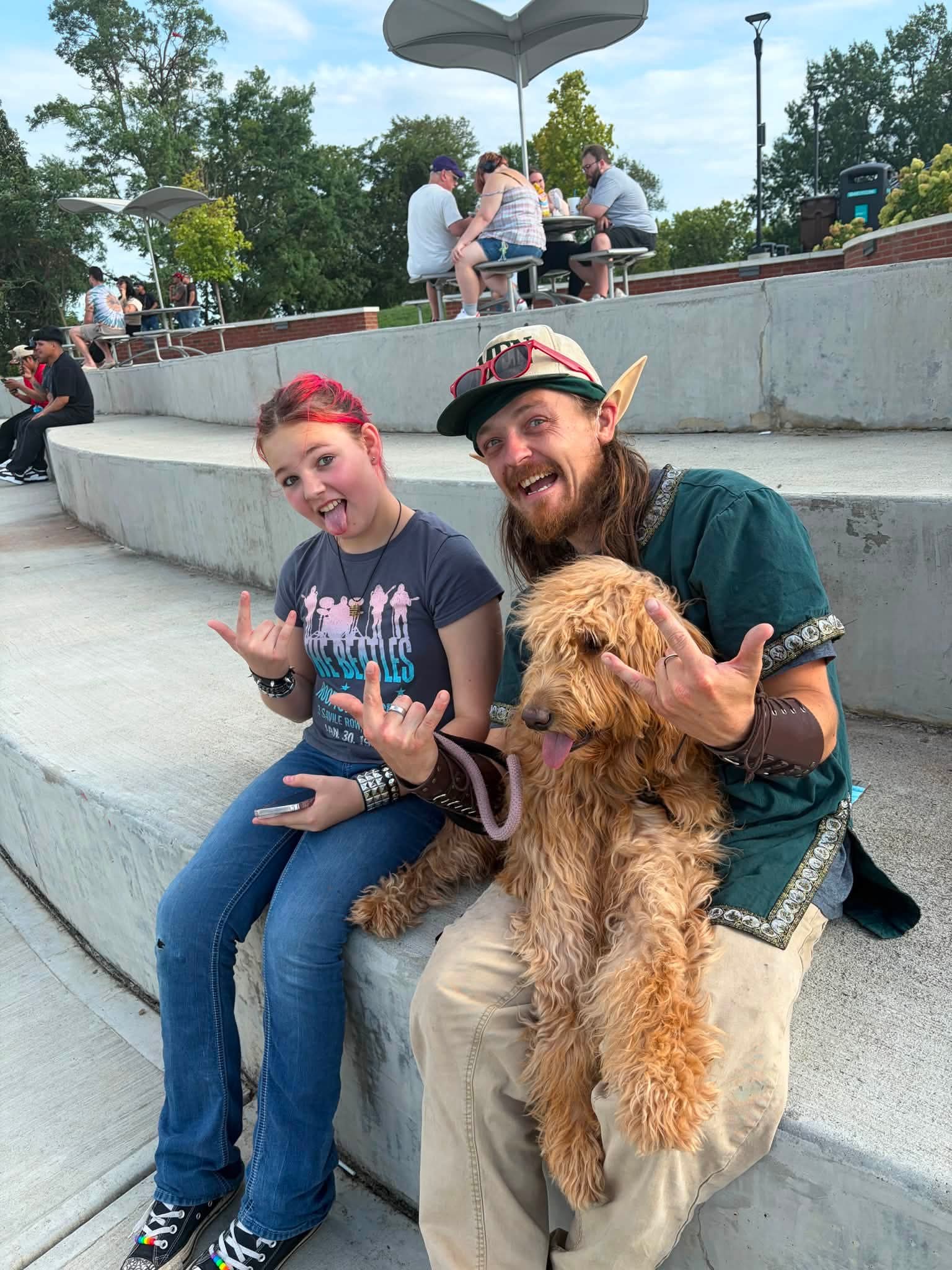 Two people and a dog sitting on a concrete step outdoors, smiling and making hand gestures, with people and trees in background.