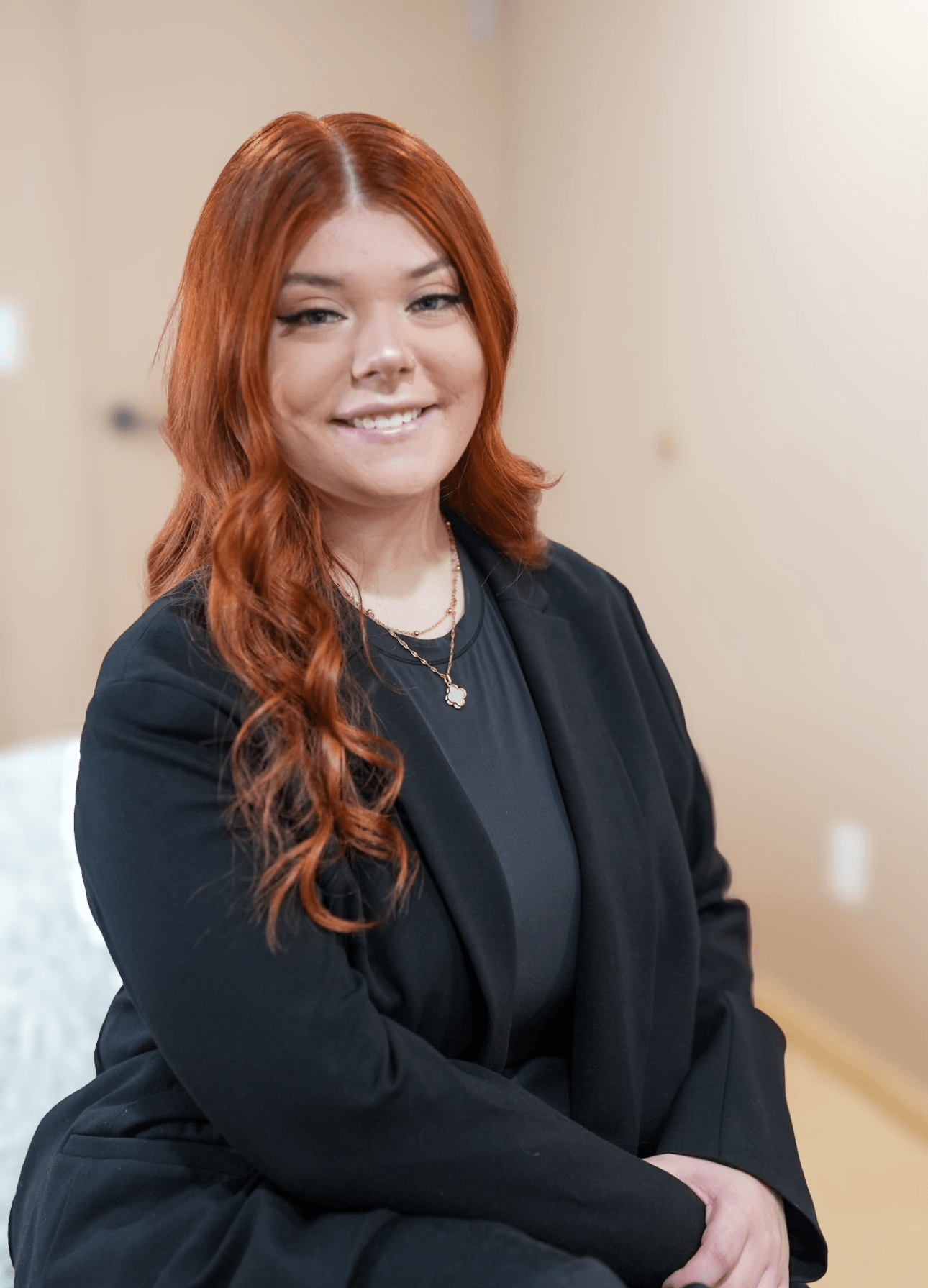 Young woman with long, wavy red hair smiling, wearing a black blazer and necklace, standing indoors.