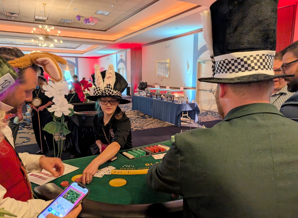 People in costumes playing a card game at a decorated indoor event with tables and colorful lighting.