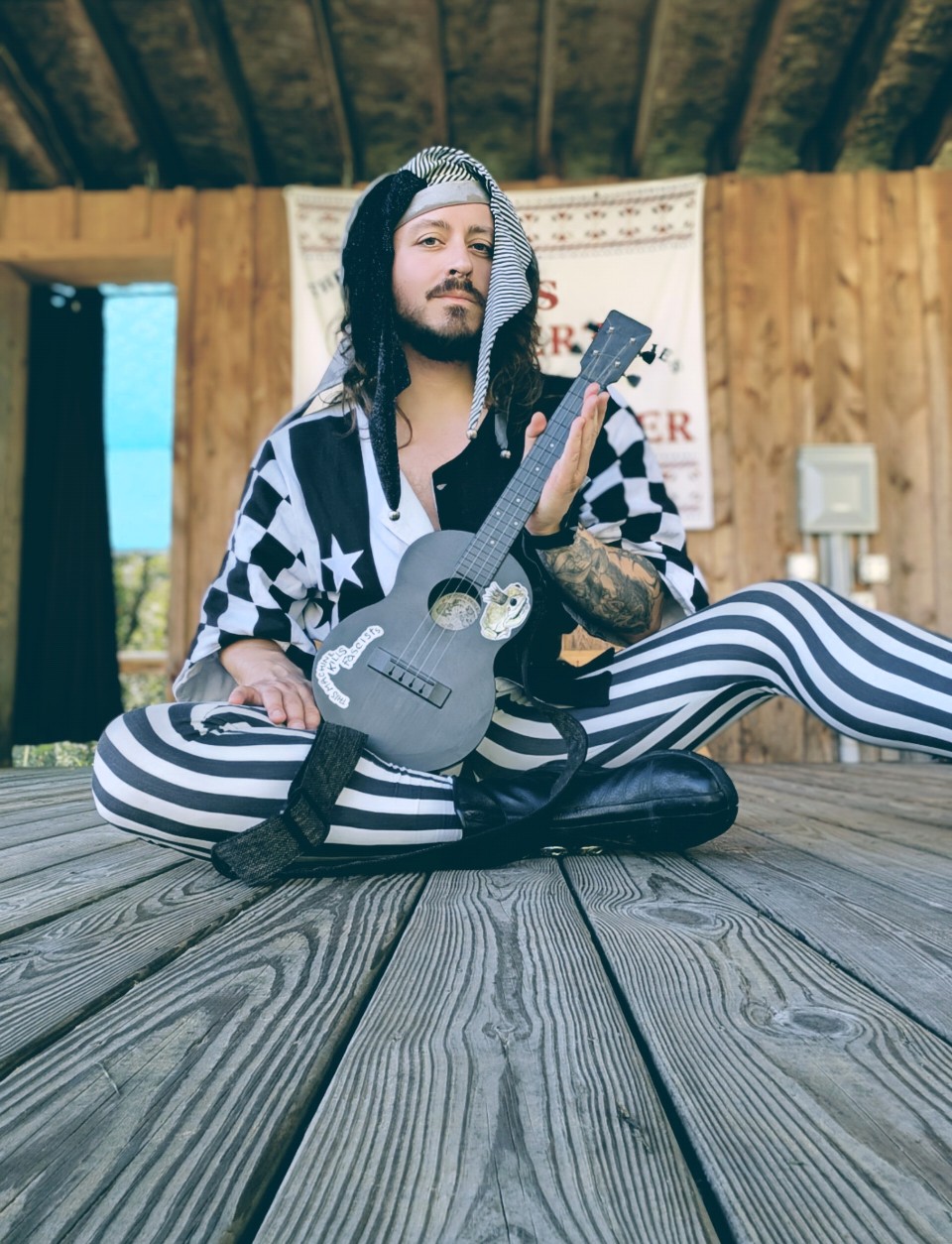 Person sitting cross-legged on wooden floor holding a guitar, wearing striped pants and a checkered shirt, with a wooden wall background.
