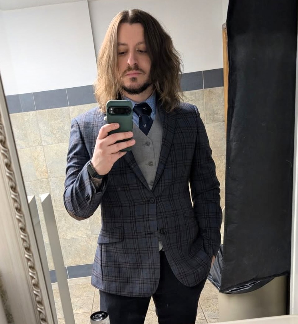 Person with shoulder-length hair in a checkered blazer taking a mirror selfie in a room with tiled walls.