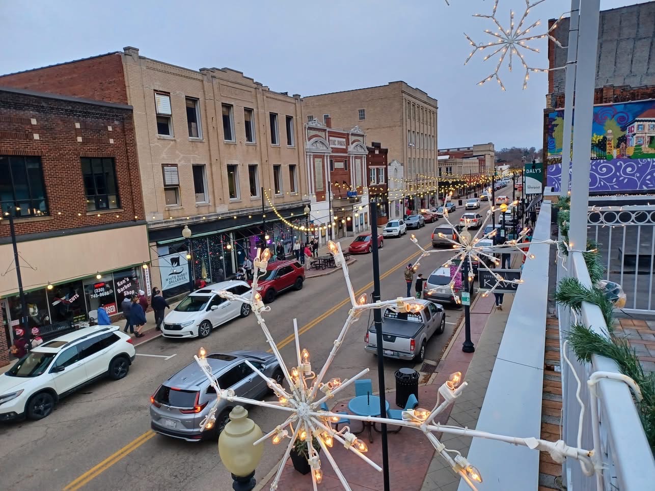 Street view with parked cars, buildings, and holiday decorations, including snowflake-shaped lights, along a busy downtown street.