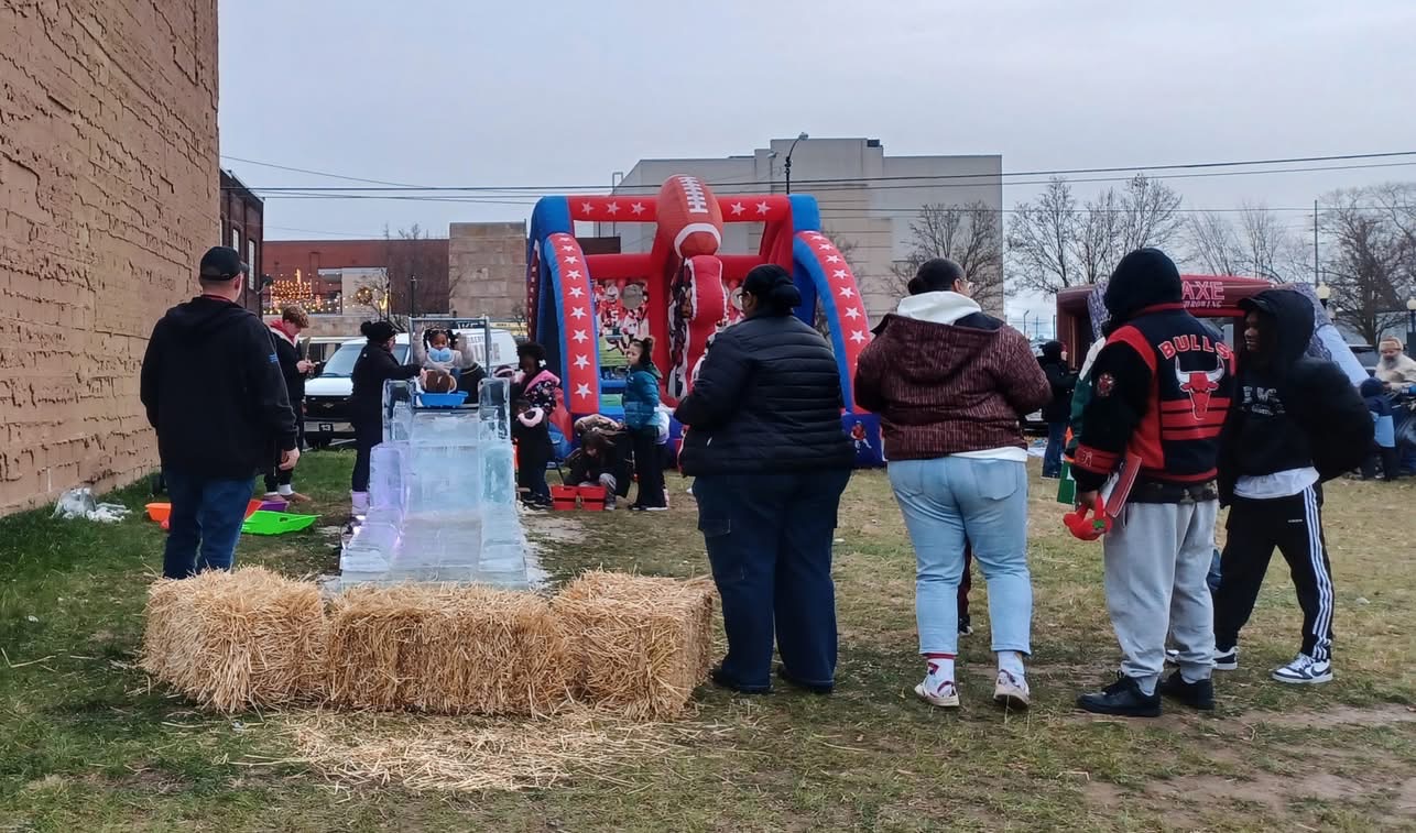 Group of people gathered around a red and blue inflatable slide outdoors, with hay bail and a clear plastic slide nearby.