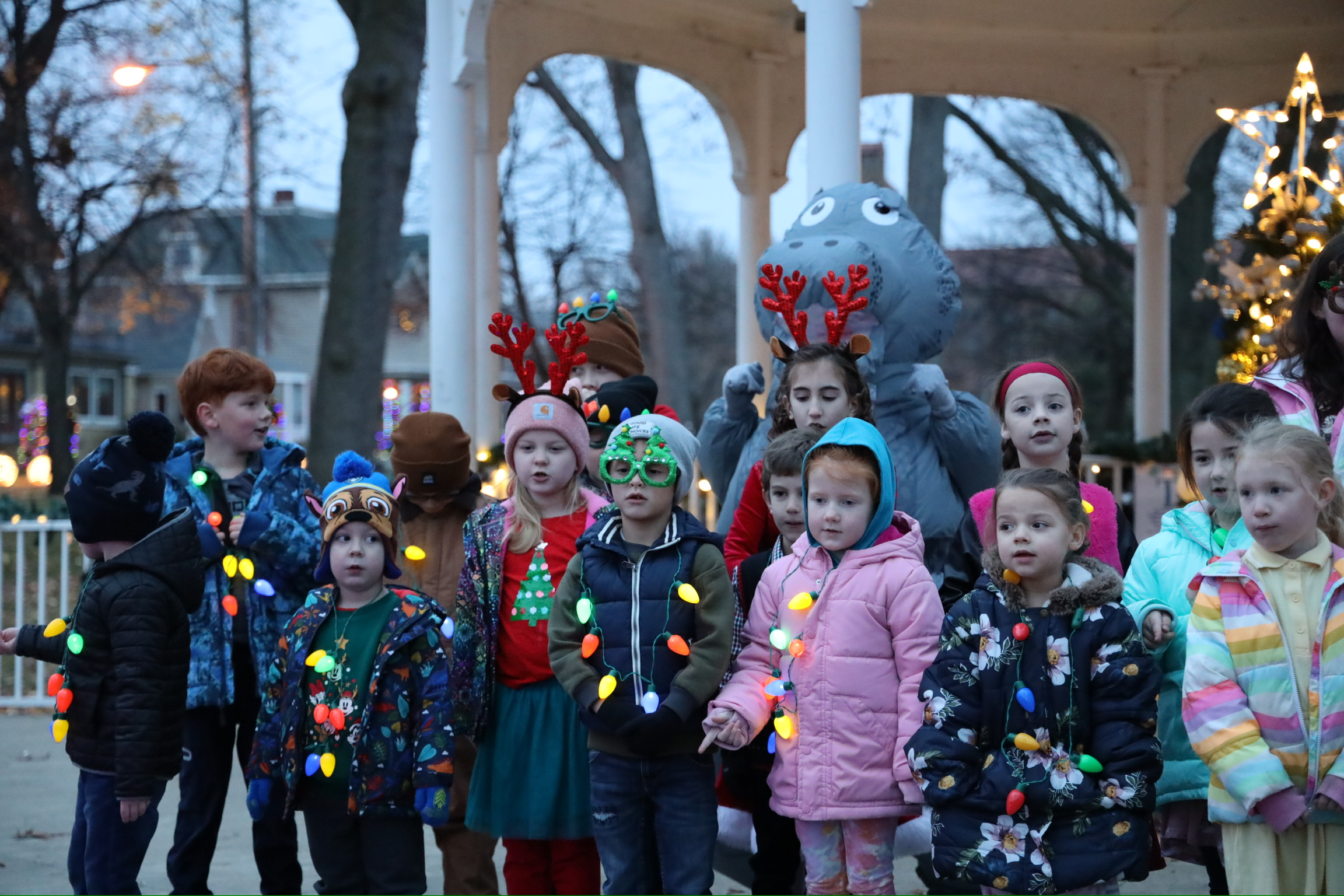 Group of children wearing winter clothes and reindeer headbands, standing outdoors near a decorated Christmas tree and a mascot, during daytime.