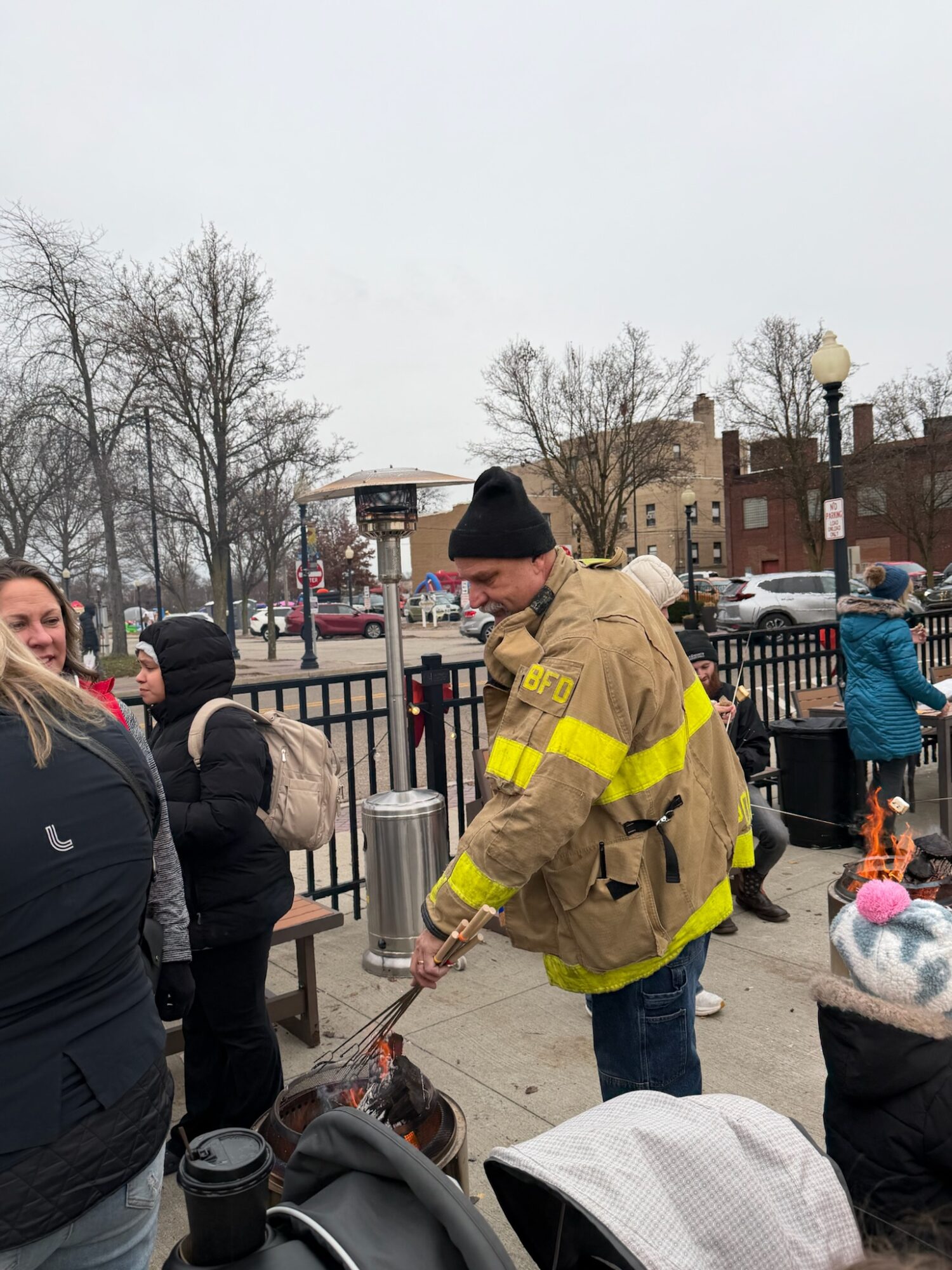 Firefighter in uniform with yellow reflective stripes interacts with children outdoors, with trees and buildings in background.