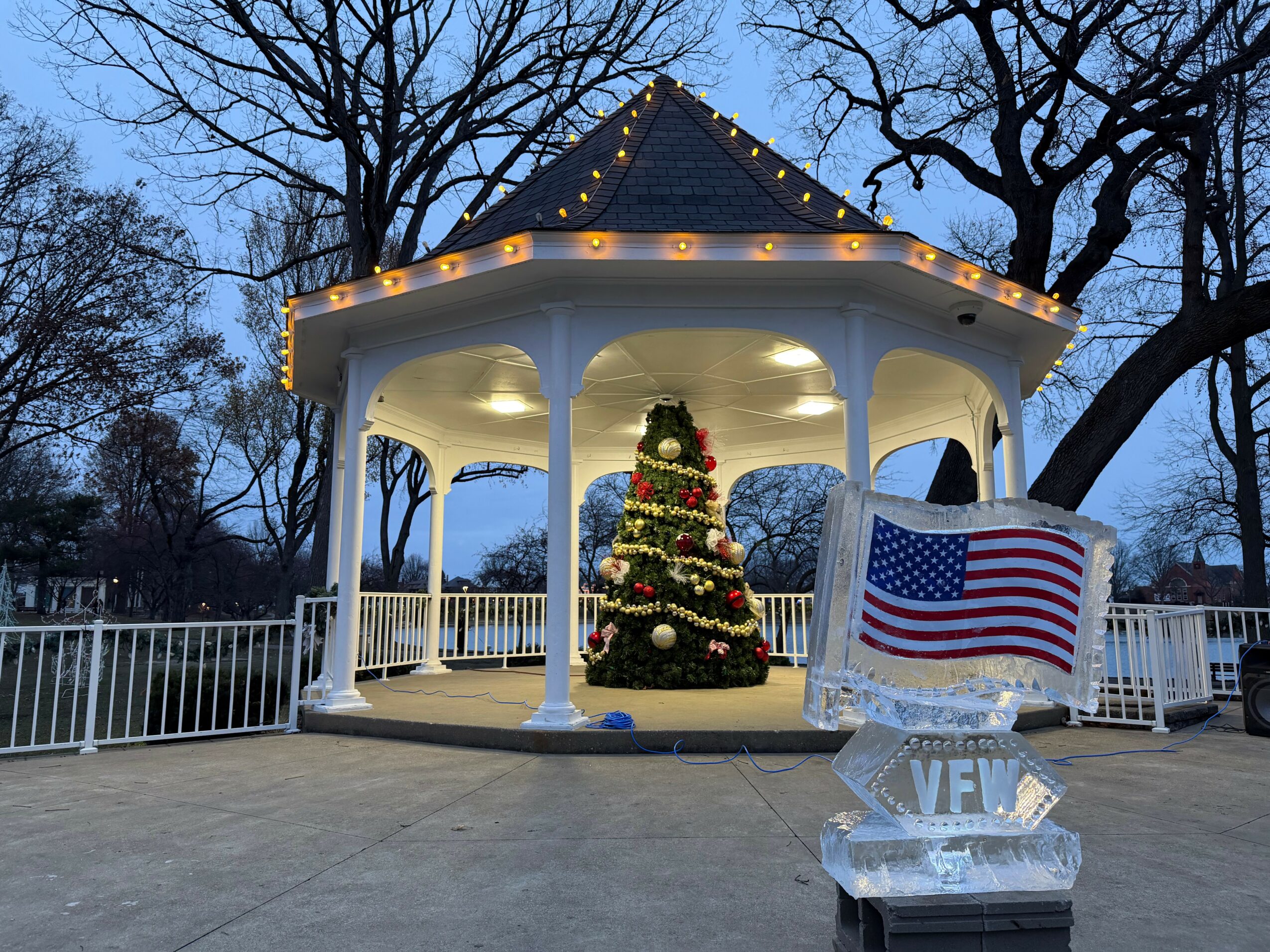 A decorated Christmas tree inside a white gazebo with string lights, American flag, and ice sculpture, outdoors at dusk.