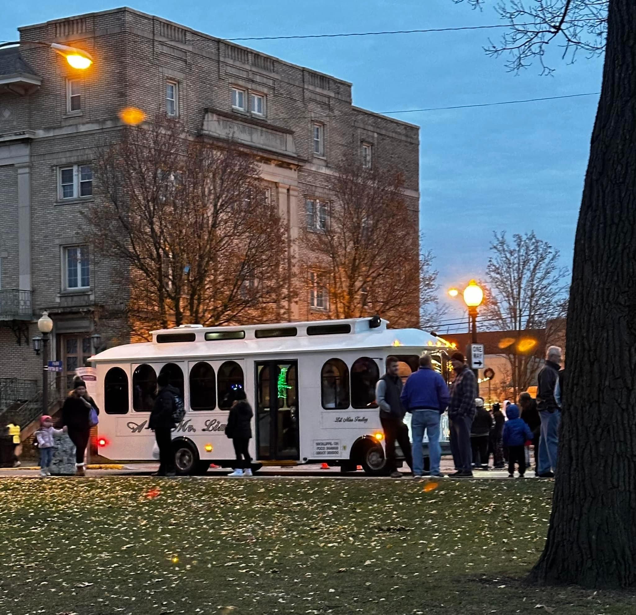 People boarding a white trolley bus on a city street during evening, with trees and a building in the background.