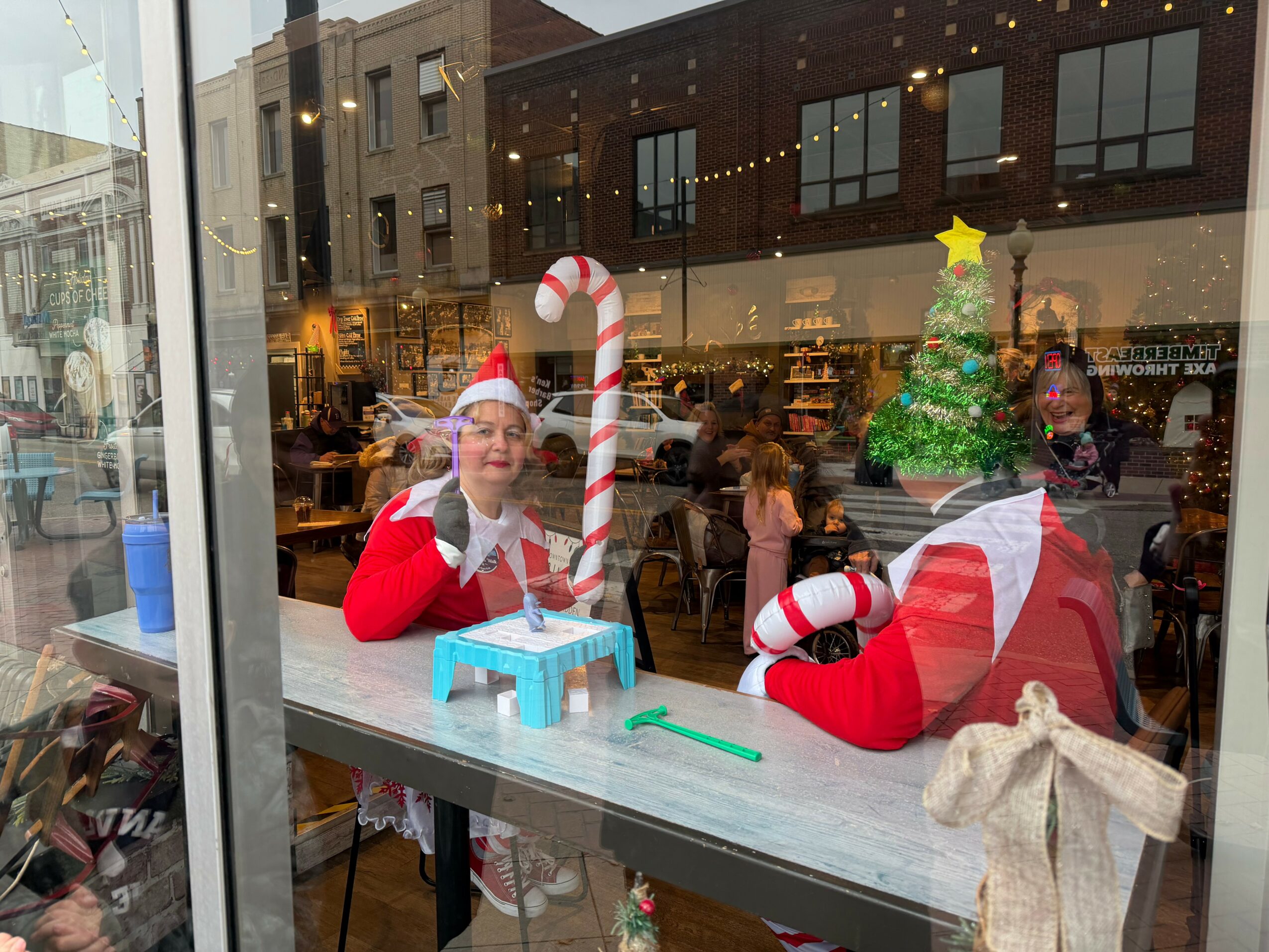Two women dressed in Christmas costumes with Santa hats, sitting at a window display with holiday decorations, including a small Christmas tree and a candy cane, inside a store. Reflection of street scene outside visible.