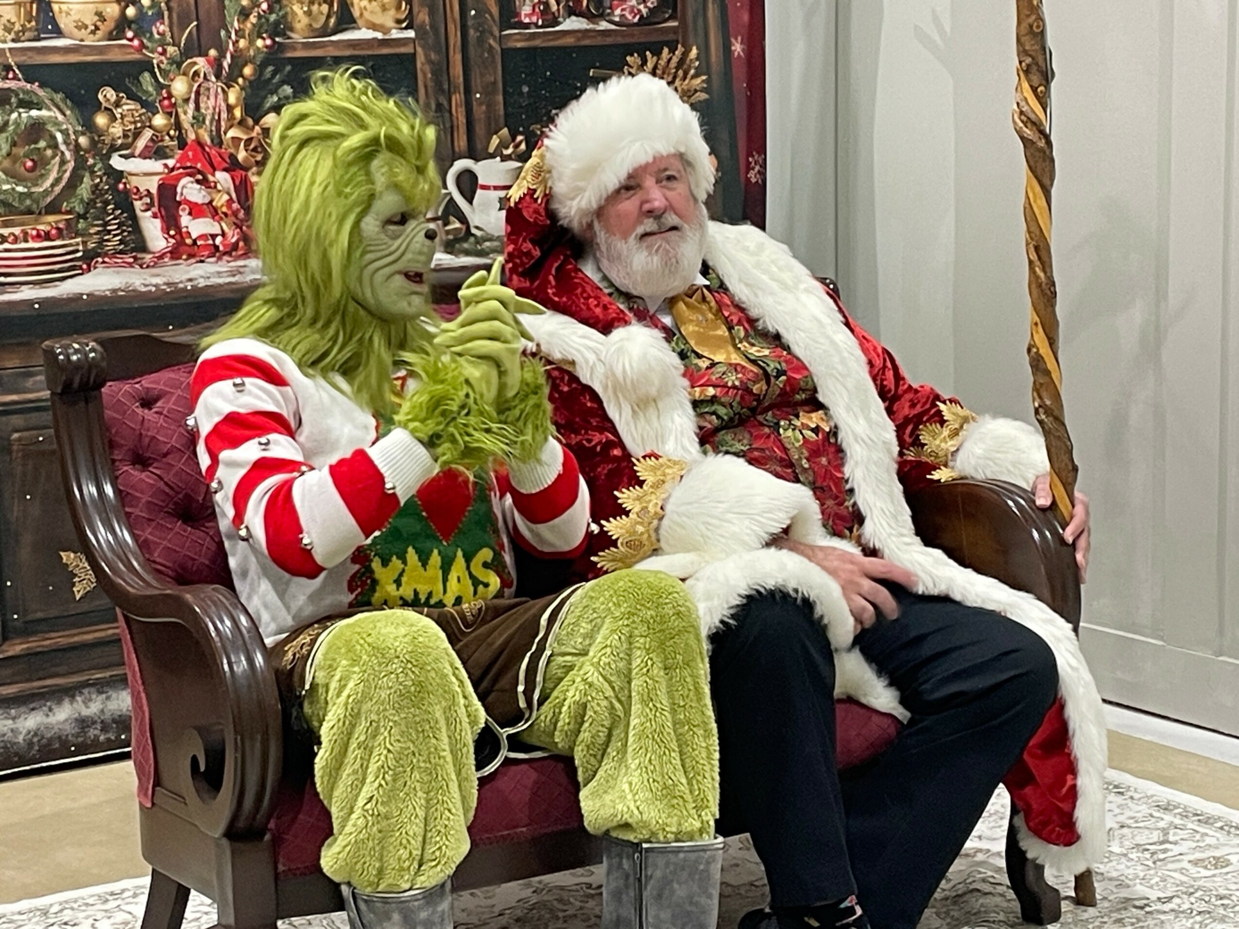 Person dressed as the Grinch and Santa Claus sitting on a red velvet bench, with festive decorations in the background.