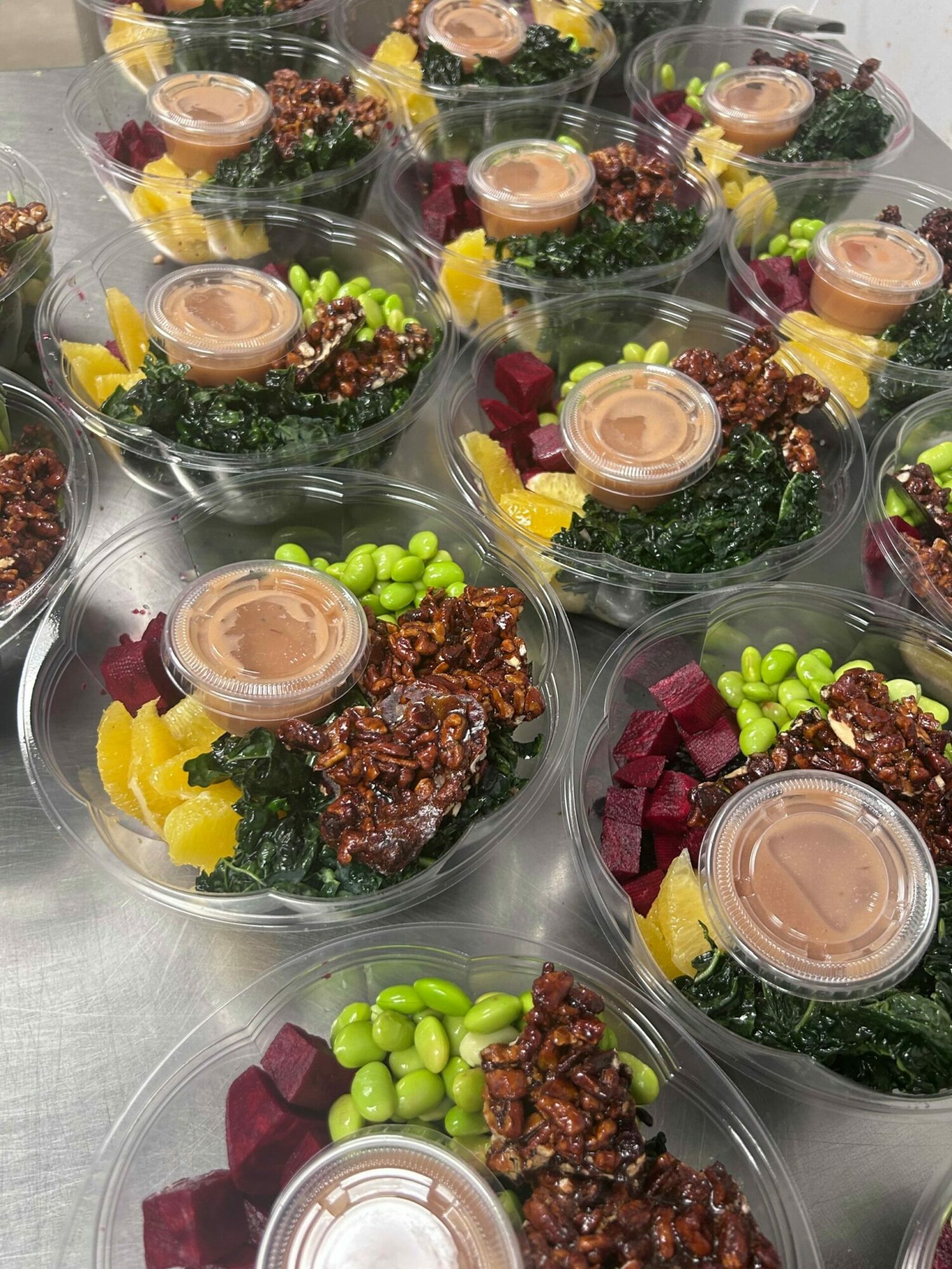 Multiple clear bowls with salads containing greens, beets, edamame, and dressing, arranged on a metallic surface.