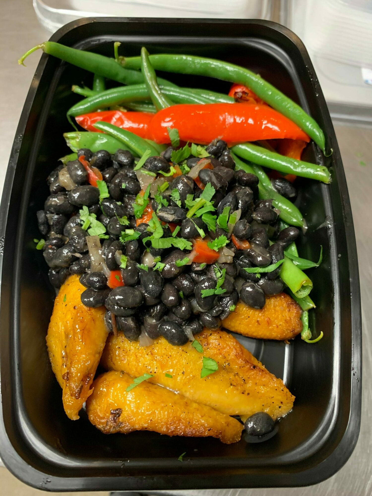 Black beans, green beans, red bell pepper, fried plantains, and chopped herbs in a black container.