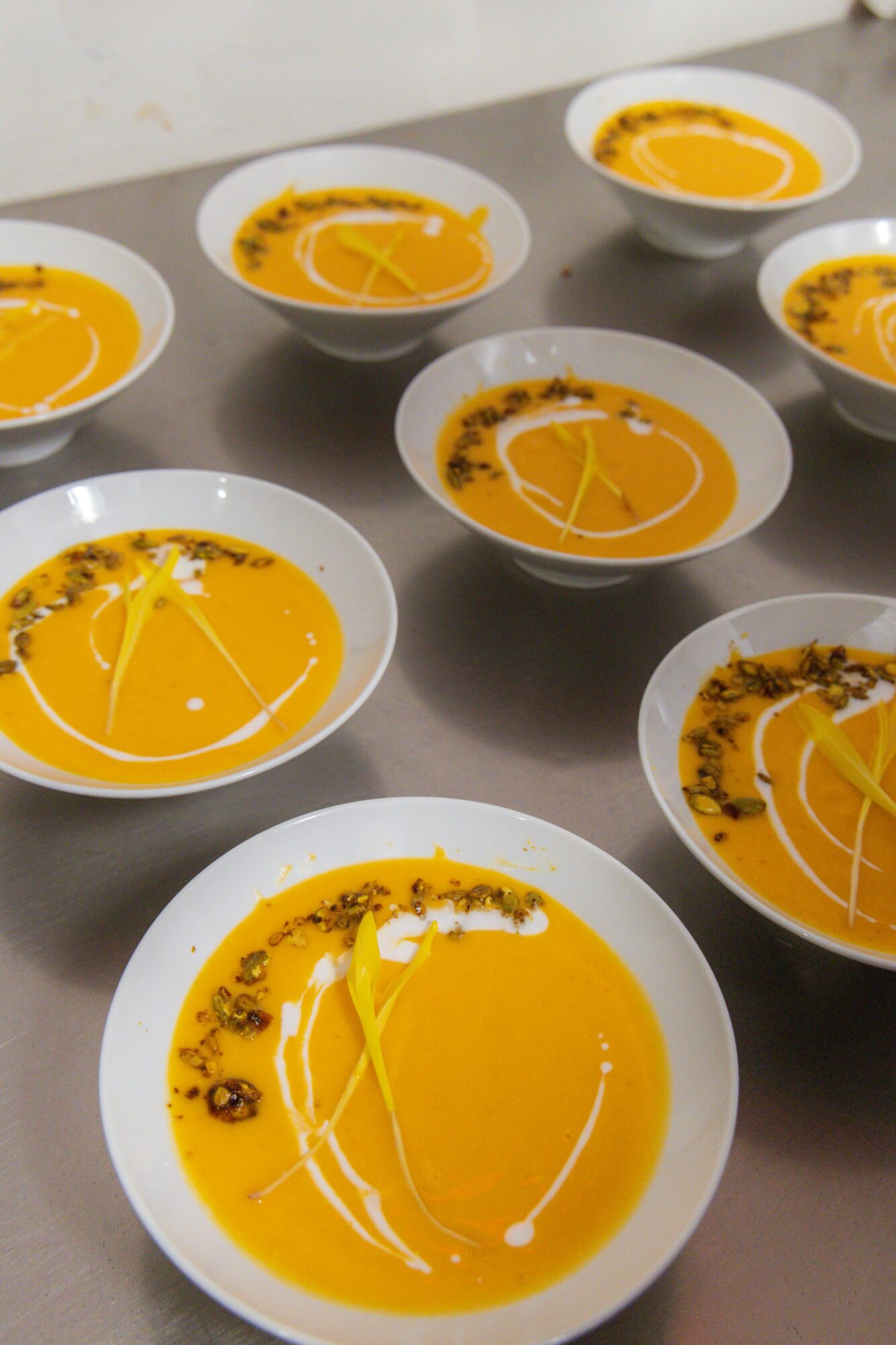 Multiple white bowls filled with orange soup, garnished with herbs and drizzled with cream, arranged on a gray surface.