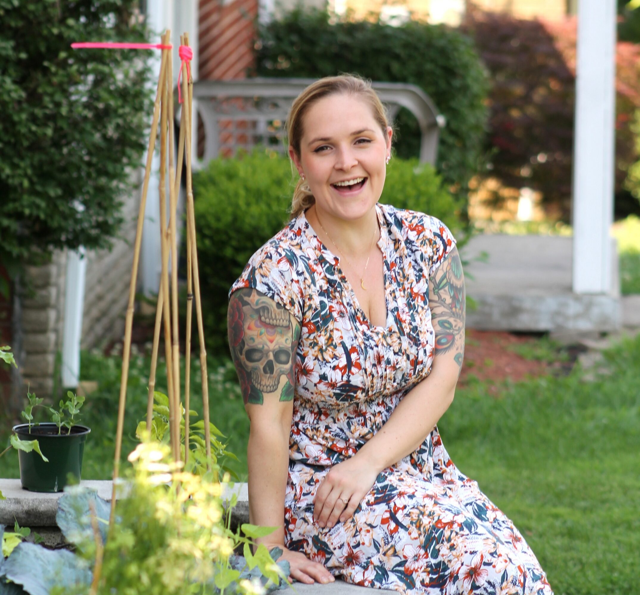 Smiling woman with tattoos on arms sitting outdoors in a garden, wearing a floral dress.