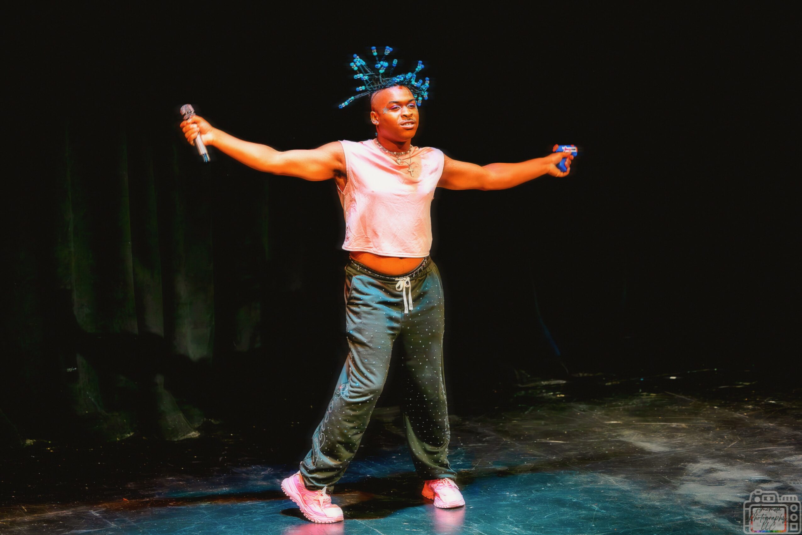 Person standing on stage with arms outstretched, wearing casual clothes and a headpiece, holding microphones in both hands.