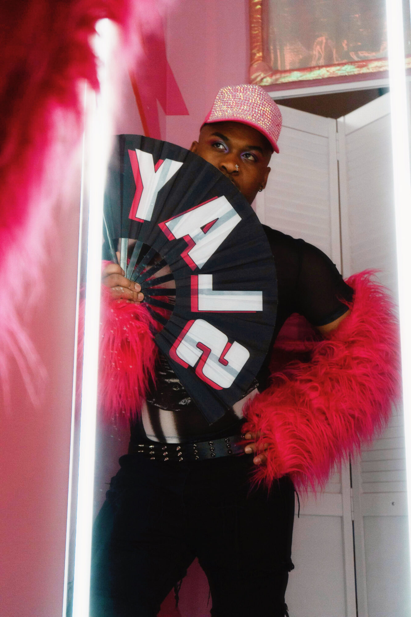 Person wearing a pink hat and red feathered coat, holding a fan, standing indoors near a mirror and white door.