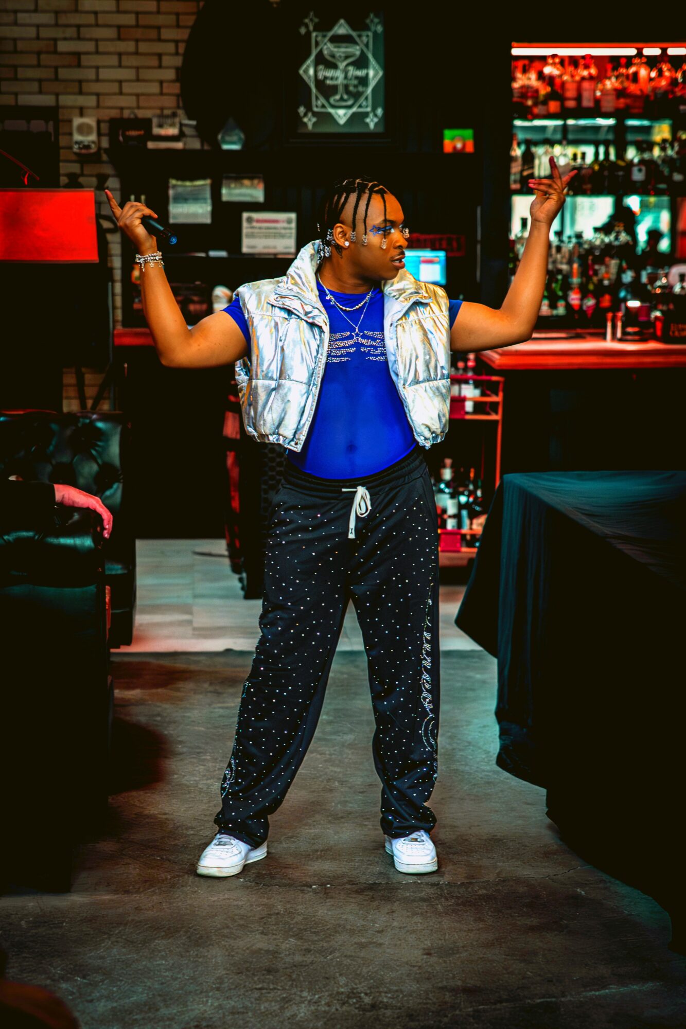 Person with glasses and braided hair stands with arms raised in a bar or club setting.