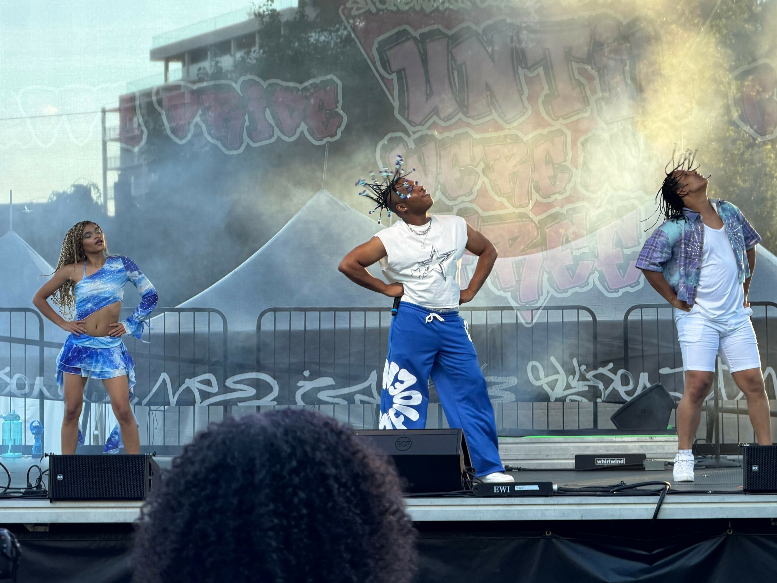 Three performers on stage with smoke effects, wearing casual outfits, dancing outdoors during daytime.
