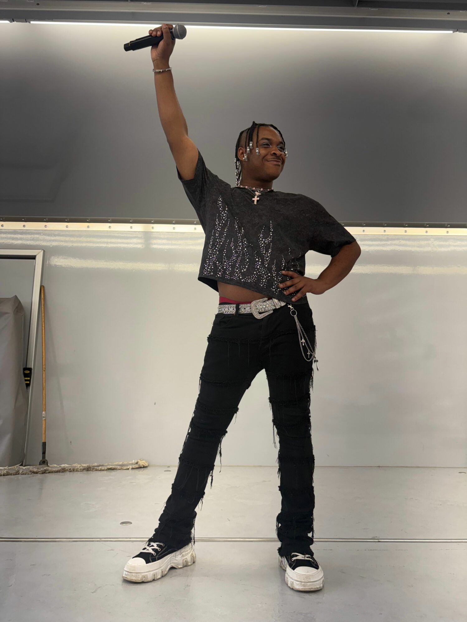 Person standing in a room, holding a microphone, with one hand on their hip, wearing a dark shirt and pants.