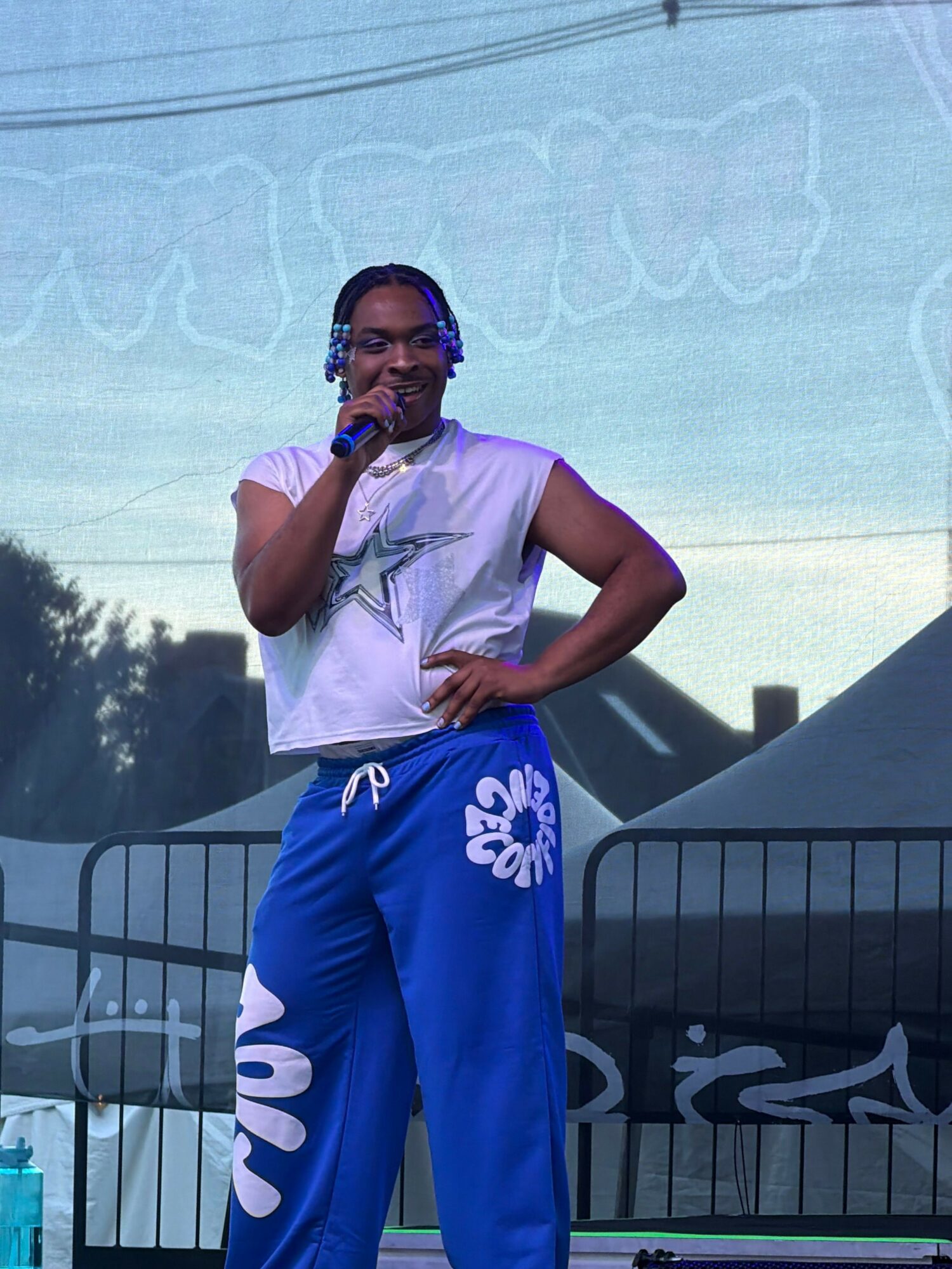 Person standing outdoors, holding a microphone, wearing a white t-shirt and blue pants, with a railing and sky in background.